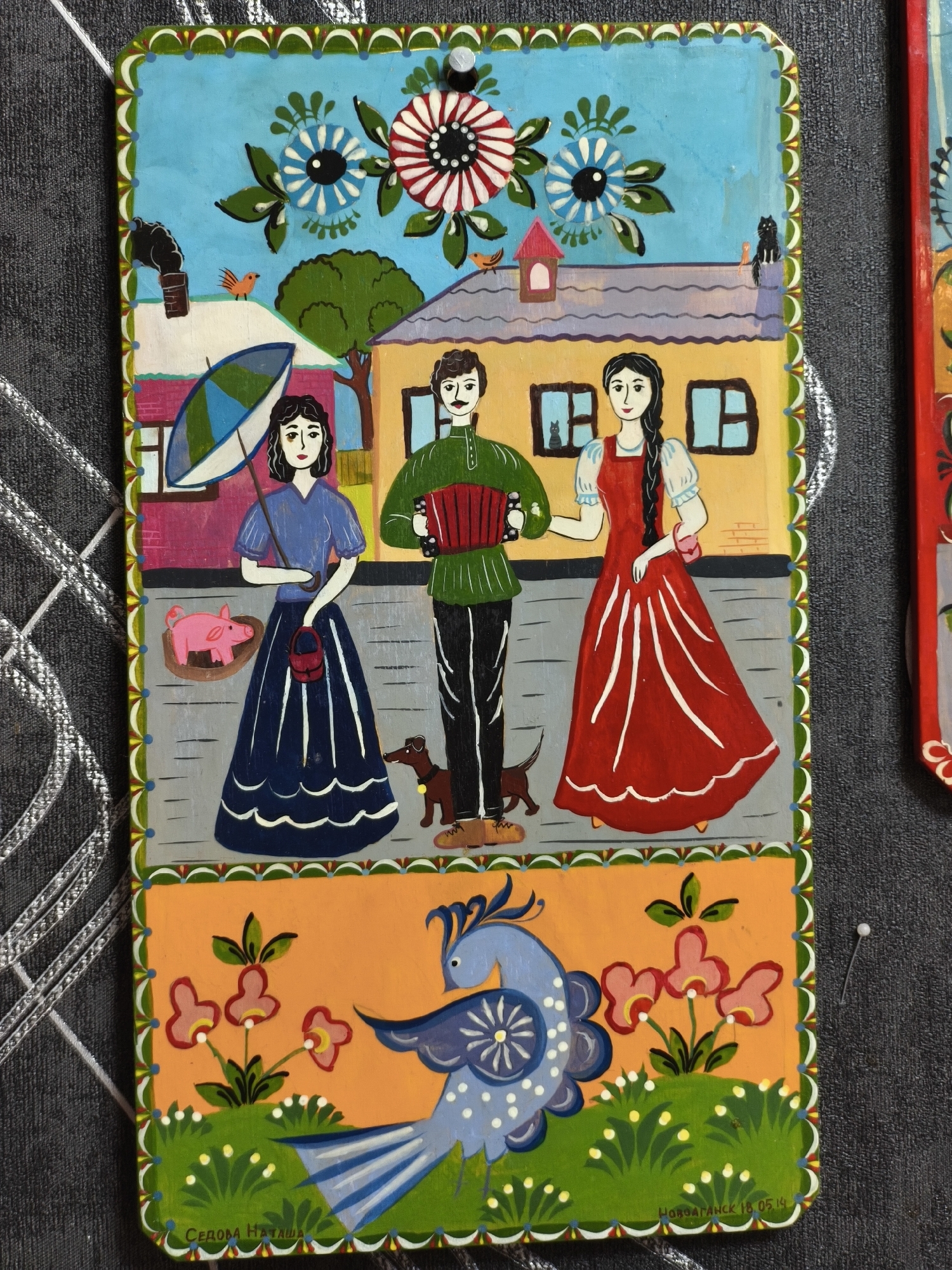 This is a vibrant folk art painting on a rectangular board, depicting a lively rural or village scene above and a stylized natural motif below. The artwork is characterized by bold outlines, flat colors, and simplified forms, typical of naive art.
The upper section of the painting features a bright blue sky adorned with large, stylized white, blue, and red flowers and green foliage. Below, two houses stand on a gray path. The house on the left is pink/purple with a white roof, a smoking chimney, and an orange bird perched nearby. The house on the right is yellow/orange with a gray roof, a small birdhouse, and a black cat sitting on the ridge. Another black cat is visible through a window.
Three human figures stand on the path. On the left, a woman with dark hair wears a blue dress, holding a patterned blue and green umbrella and a small red handbag. Beside her, a pink pig rests in a wooden trough. In the center, a man with dark hair and a mustache wears a green shirt and black pants, playing a red accordion. A small brown dog stands near his feet. On the right, a woman with dark hair in a long braid wears a long red dress with white sleeves. The figures have serene, slightly smiling expressions.
The lower section of the painting has an orange background. A large, stylized blue bird with intricate white and blue patterns on its body and tail feathers dominates this area. It is surrounded by green grassy patches dotted with white flowers and several pink flowers with yellow centers.
The painting appears to be mounted on a dark, textured surface with a white-headed pin visible in the top right, holding the artwork in place.
Two lines of Cyrillic text are visible at the bottom of the artwork. On the left, it reads "СЕДОВА НАТАЛЬЯ" (SEDOWA NATALYA), likely the artist's name. On the right, it says "НОВОГРАНСК 18.05.14" (NOVOGRANCK 18.05.14), indicating a location and the date May 18, 2014. The bright colors and detailed imagery suggest a joyful and idyllic portrayal of rural life and nature during daytime.