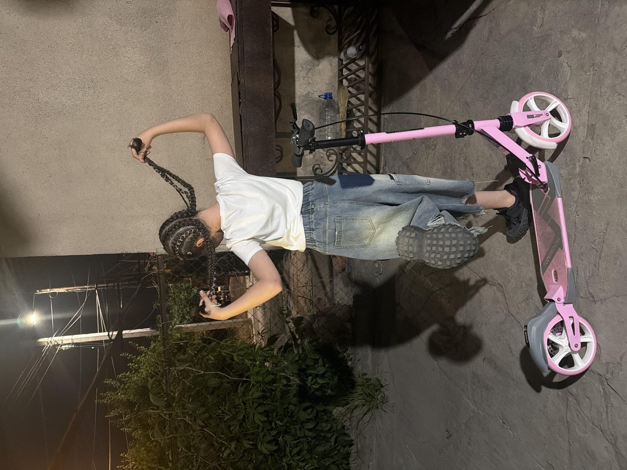 This nighttime image, set in an outdoor area in Yerevan, Armenia, features a person standing next to a pink kick scooter. The individual, viewed from the back and side, has numerous dark braided hairstyles and is holding a section of their braids with their right hand. They are dressed in a white short-sleeved t-shirt, loose-fitting, light blue distressed denim pants, and dark sneakers. Their left foot is positioned on the scooter's deck.
The kick scooter is predominantly pink with white wheels and gray elements. The brand name "TOPENOUN" is visible on its deck.
The background shows a light-colored wall on the left, a dark metal fence or gate with intricate patterns, and dense green bushes behind the fence. The ground is a dark, textured paved surface. Artificial light sources are visible, including a bright light in the upper left, casting shadows and illuminating parts of the scene, confirming the nighttime setting. A plastic bottle can be seen near the fence behind the scooter's handlebars.