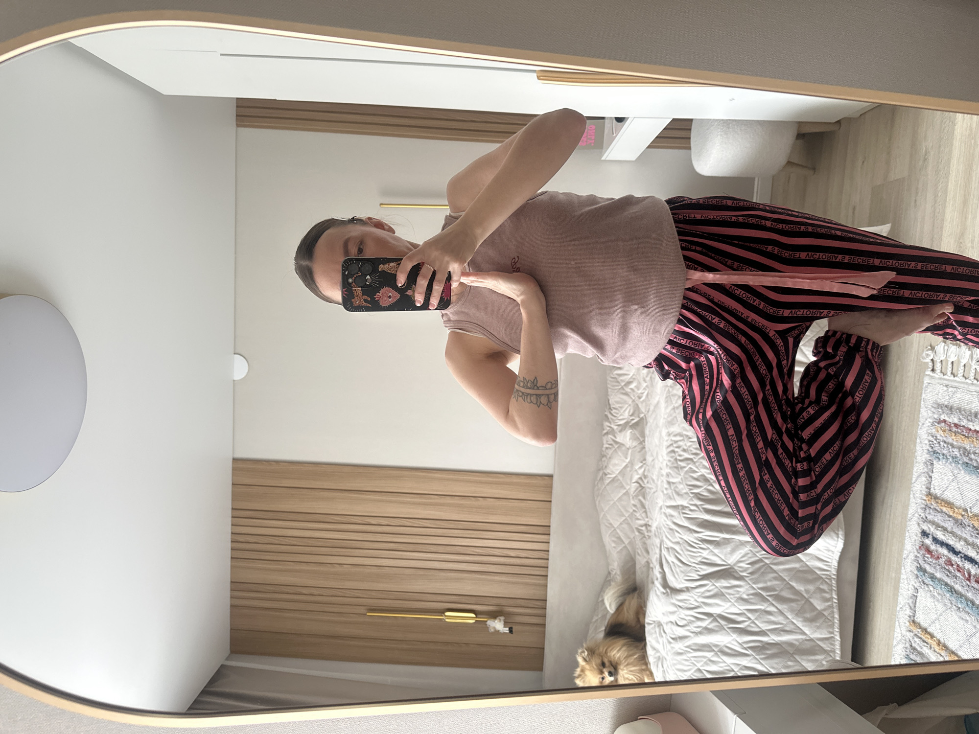 A person is seen in a mirror selfie. The person has brown hair, wears a mauve tank top and pink and black striped pajamas. The location appears to be a bedroom with white walls, a wooden slat wall, a bed with a white quilted blanket, and a small dog on the bed. There is a white table with a white chair or stool. There is a mirror with a gold frame. The location is not specified, and there is no indication of the city of Votkinsk, Russia, being involved.