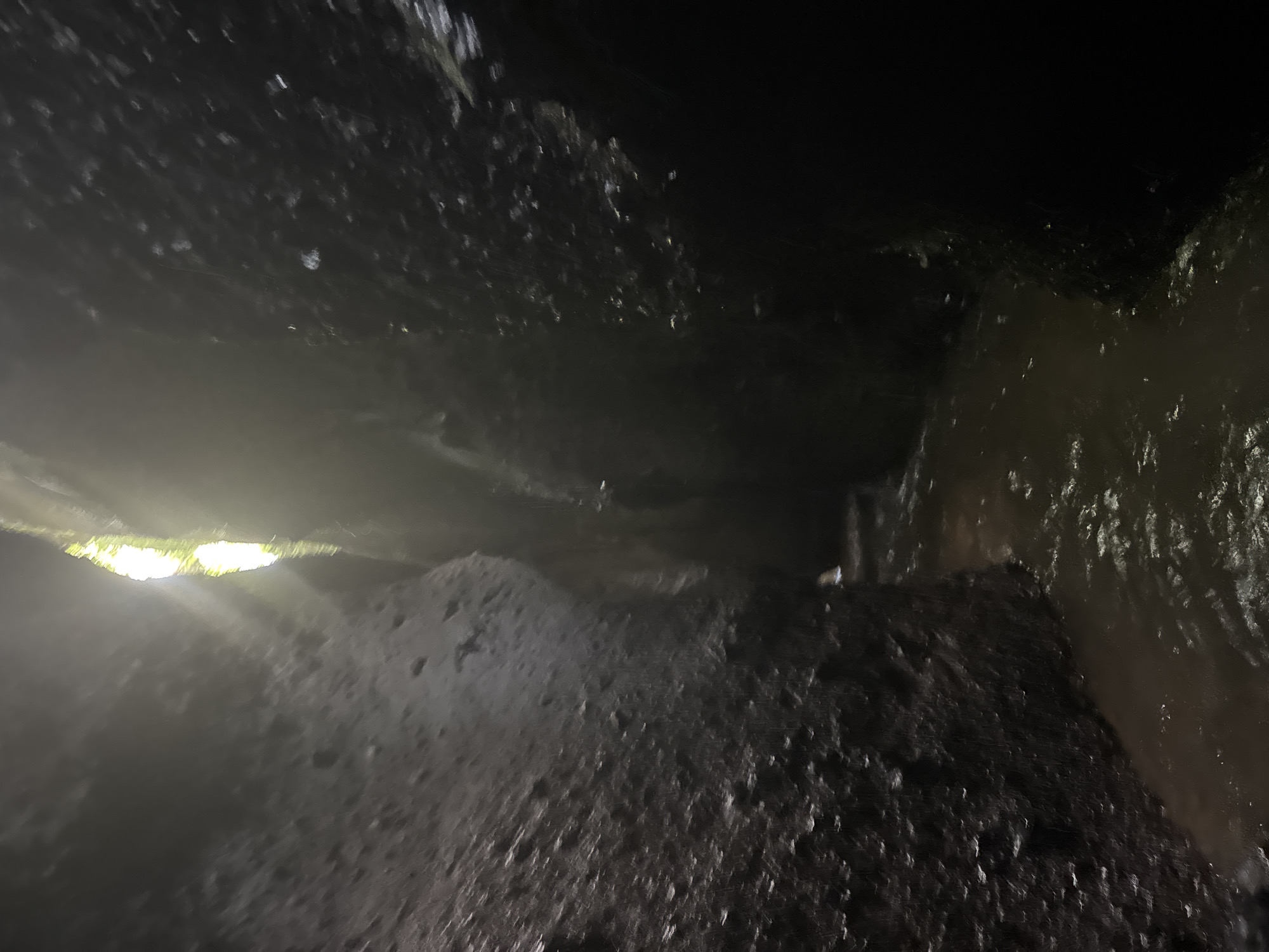 This is a photograph of a narrow canyon with water flowing through it. The canyon walls are dark and rocky, with some vegetation visible at the top where sunlight is filtering through. The water is shallow and appears to be moving. The overall impression is one of a natural, secluded place. The location is identified as Tembuku, Indonesia, which is a sub-district in the Bangli Regency of Bali, known for its natural beauty and tourist attractions, including waterfalls and natural pools like the one depicted here.