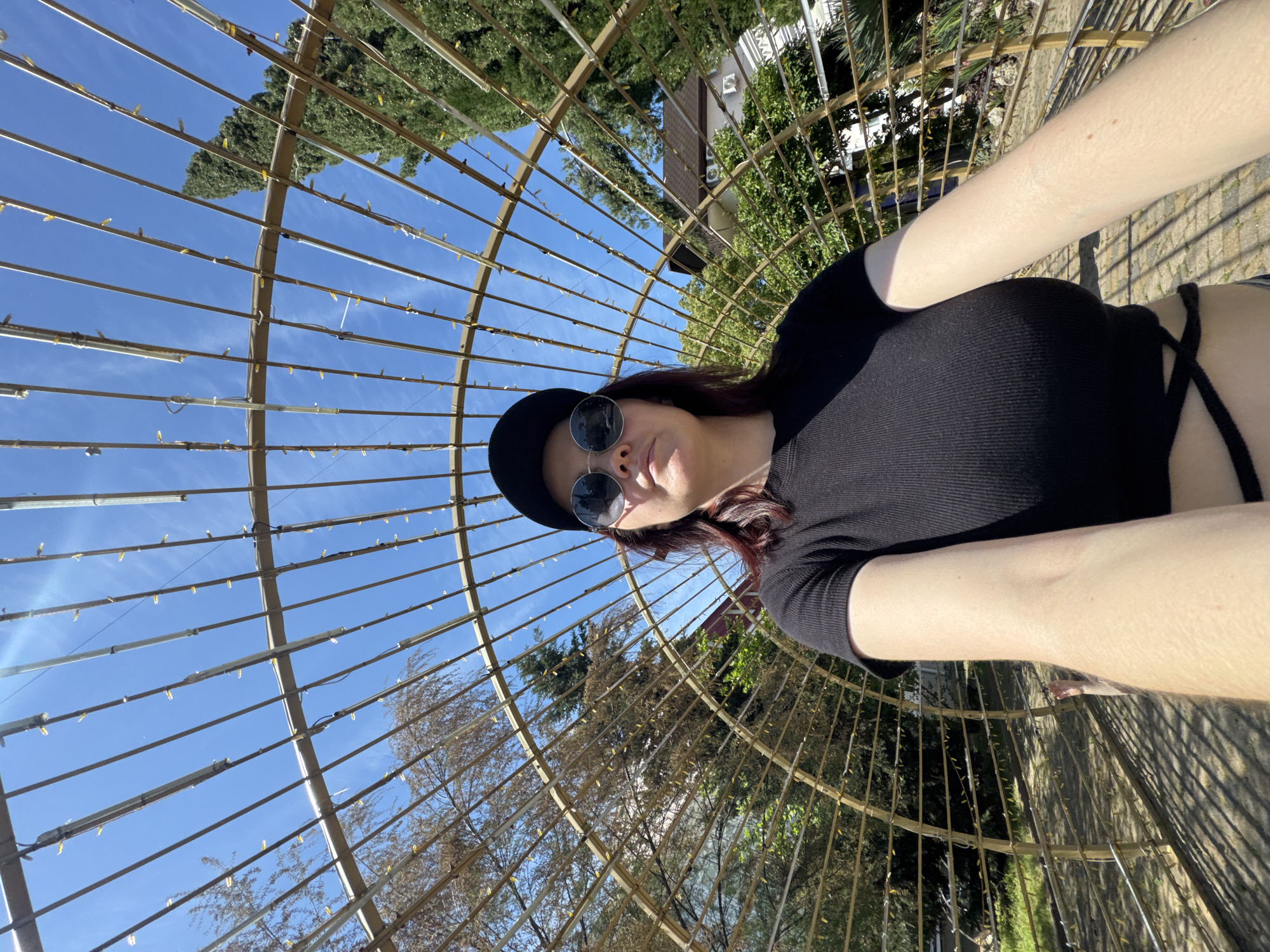 A young adult woman poses for a selfie inside a large, spherical metal structure with many thin, parallel bars. The structure is outdoors, with trees and buildings visible in the background. The sky is blue. The woman is wearing a black baseball cap, sunglasses, and a black top. The photo appears to have been taken with a wide-angle lens, causing some distortion. The image location is likely Sochi, Russia.