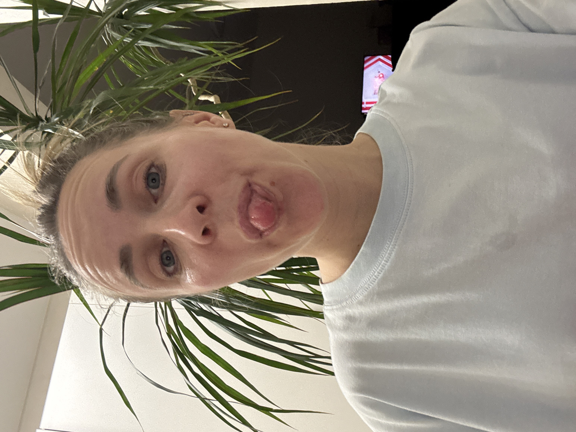 A close-up selfie shows a person, likely a woman, with a playful expression, her tongue slightly out. Her face, featuring blue eyes and light hair pulled back, is viewed from a low angle and is partially framed by the long, green leaves of a large indoor plant. She is wearing a light-colored, possibly white or light blue, t-shirt.
The setting appears to be an indoor space in Chaikovsky, Russia, possibly a living room, as a television screen is visible in the background. The screen displays a colorful image or video, featuring what looks like a cartoon character surrounded by red and white striped borders. The lighting is ambient, characteristic of an indoor environment, without strong indicators of the time of day. The overall mood conveyed by the subject's expression is casual and humorous. No discernible text is visible on the subject or within the immediate surroundings apart from indistinct elements on the television screen.
