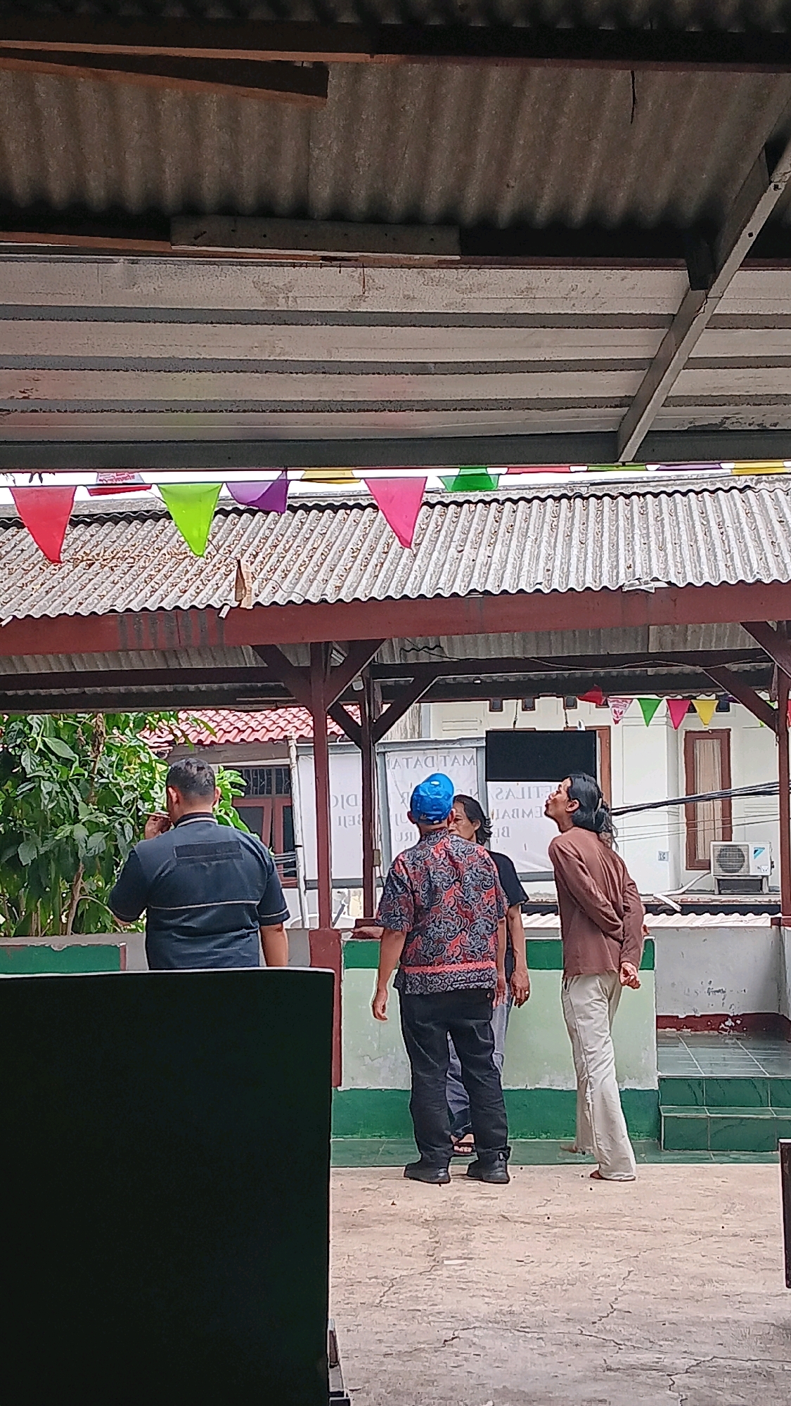 Four individuals are standing outdoors under a covered structure in Depok, Indonesia. The structure has a corrugated metal roof and is decorated with colorful triangular flags. In the foreground, a person wearing a dark blue shirt and black pants stands with their back to the camera, holding something to their mouth. To their right, another person wears a patterned shirt with red and blue designs and a blue cap. They are facing away from the camera and looking upwards. Beside them, a person in a brown long-sleeved shirt and light-colored pants is also looking upwards, with their back to the camera. Partially visible to the right of the person in the blue cap is another individual in a dark shirt. The ground is paved with concrete. In the background, there is a building with windows and an air conditioning unit. Lush green foliage is visible to the left. The scene appears to be a public or semi-public outdoor space.