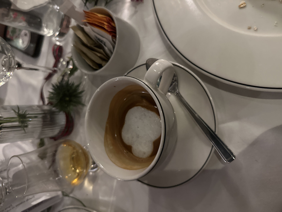 An overhead shot shows a partially consumed cappuccino in a white cup with a black rim, resting on a matching saucer with a spoon. Next to it is a white dish with dark trim and some food residue. Further left, there's a small white container filled with sugar packets. Several glassware items are also scattered around the scene, including a water glass and a small flower vase. The setting appears to be a formal dining table covered with a white tablecloth.