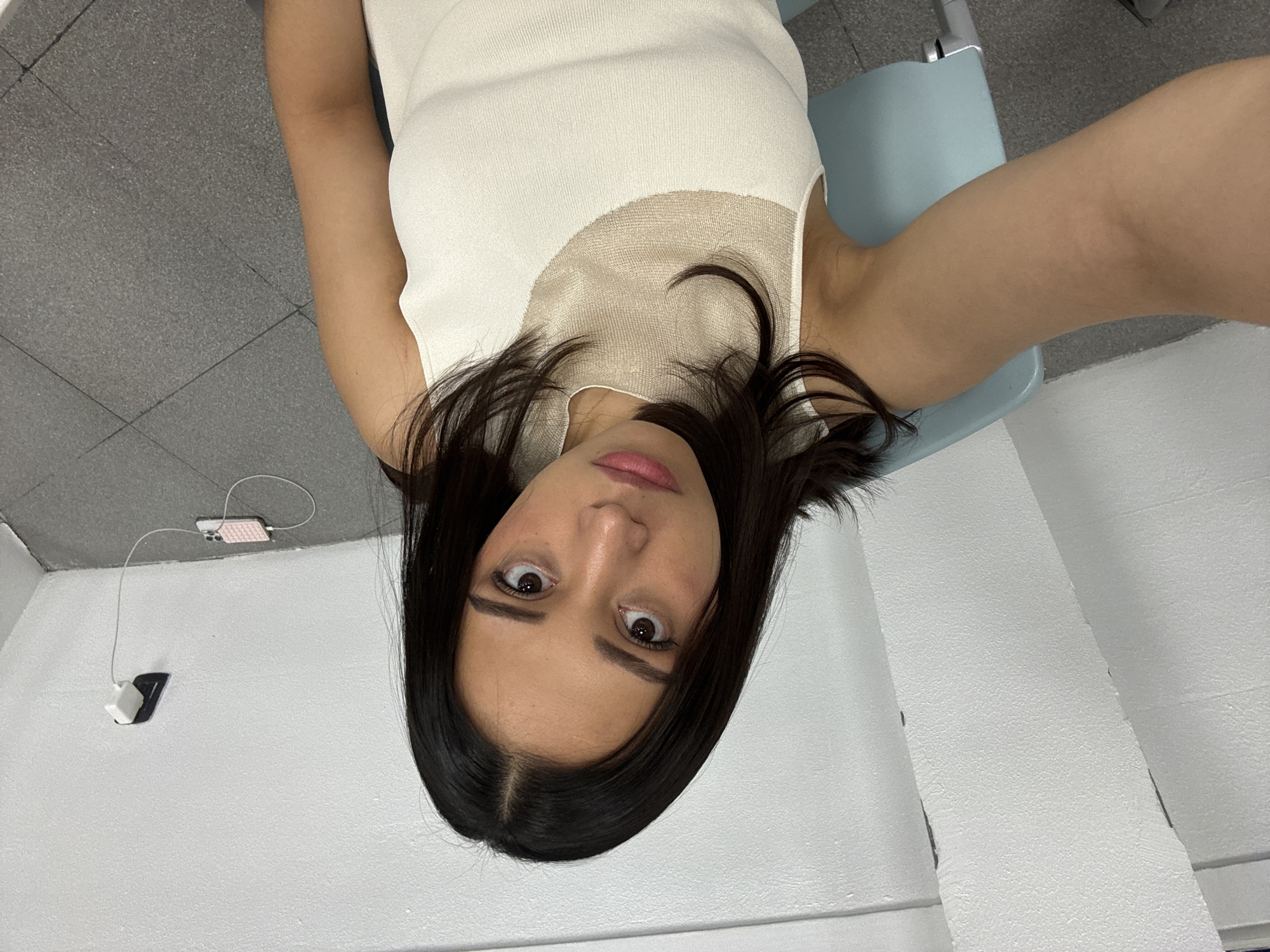 The image displays a female individual in a head-and-torso shot, captured as a selfie. She has dark, shoulder-length hair and a neutral facial expression, looking directly into the camera. She is wearing a cream-colored, sleeveless top featuring a sheer, textured mesh panel across the upper chest. A light blue chair is partially visible on the left, indicating she is seated. The background consists of plain white, textured walls, forming an interior corner on the right side of the frame. On the right wall, a black electrical outlet contains a white charging adapter. A white cable extends from this adapter to a pink smartphone resting on the dark gray tiled floor, indicating the phone is actively charging. The floor exhibits distinct grid-patterned grout lines. The environment is an unadorned indoor space, and no specific visual cues within the image provide an identifiable location within the city of Puebla, Mexico.