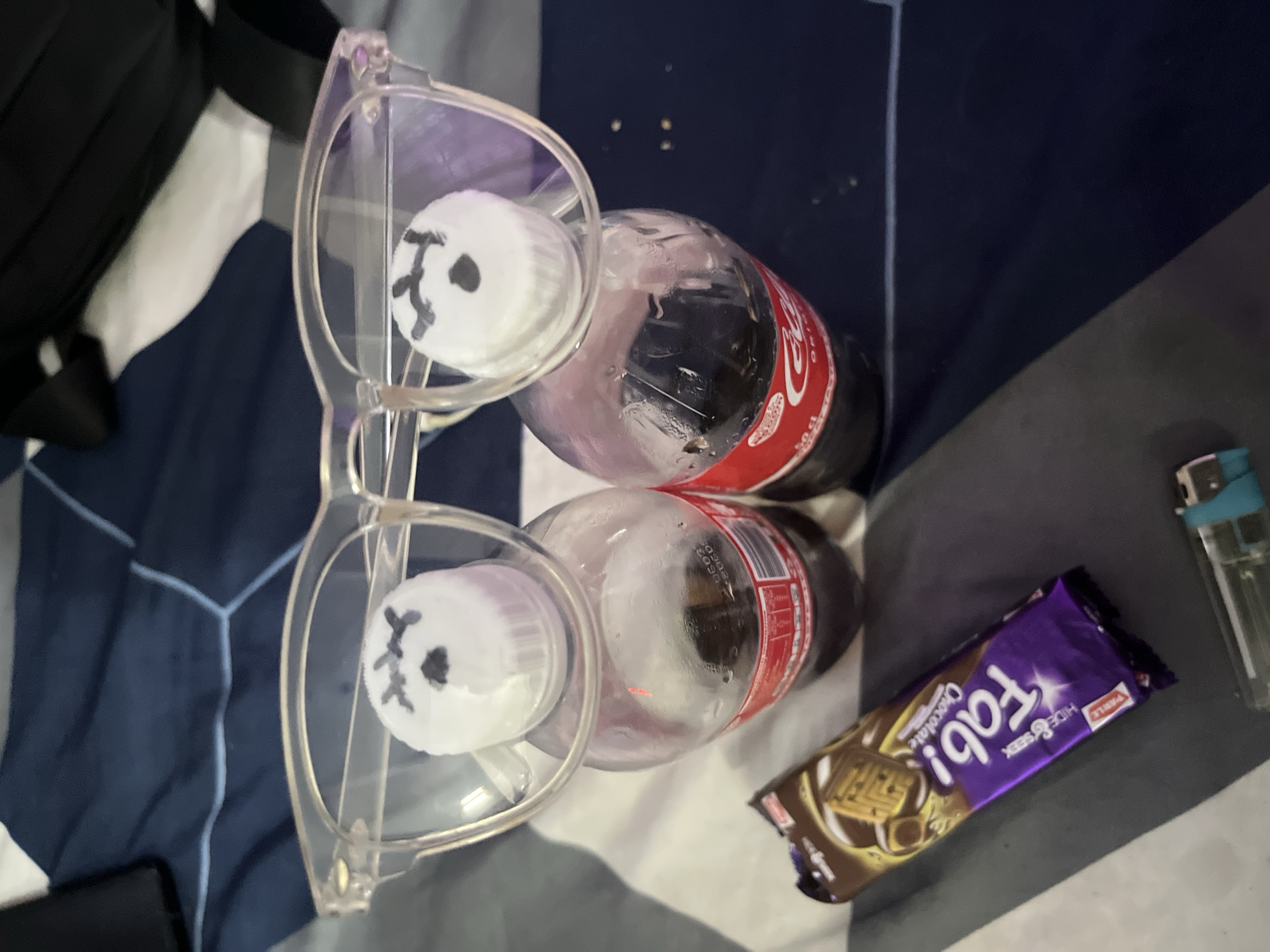 The image depicts two Coca-Cola bottles lying on a bed with a white and blue patterned sheet. The bottles are partially obscured by a pair of transparent eyeglasses. The caps of the bottles have drawings of faces on them. To the right of the bottles are a purple and brown chocolate biscuit package and a lighter. A black backpack is partially visible on the left side of the image. The scene is indoors. The location is Lokoja, Nigeria is not discernible from the image.