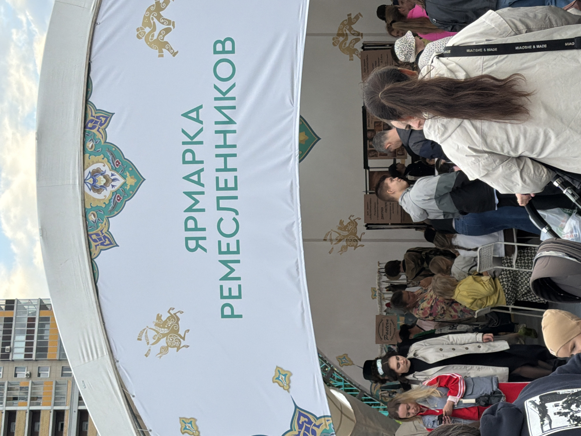 The image depicts an outdoor marketplace under a large white tent adorned with decorative borders featuring intricate patterns and golden illustrations of figures on horseback and wrestlers. The tent's canopy is partially visible, suggesting an event or fair. Dominating the foreground is a crowd of people, with a woman in a beige hooded jacket facing away from the camera being a prominent subject. She has a black strap with white lettering across her shoulder. To her left, a young person in a grey and black jacket is looking left, possibly interacting with someone off-camera. Further left, a woman in a red jacket and blue jeans is holding a mobile phone. Several other individuals are visible in the mid-ground, some standing and some seated at tables, suggesting stalls or vendors. The overall atmosphere appears to be lively and social, characteristic of a public gathering. The background shows a building with multiple floors and balconies, indicating an urban setting. The sky above is partly cloudy, suggesting daytime.