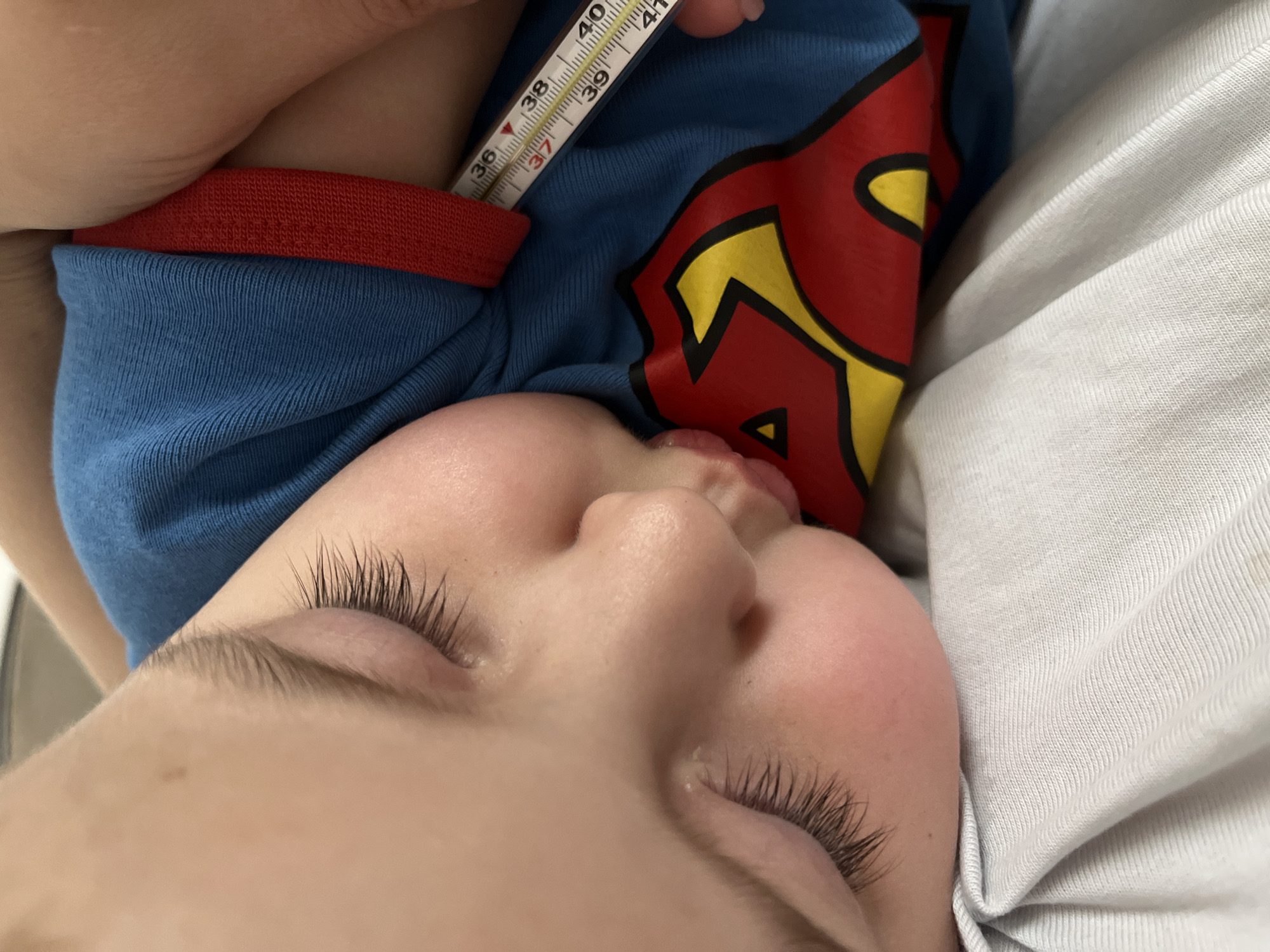 A close-up, eye-level shot shows a sleeping child lying on their side, with a thermometer tucked under their shirt. The child's face, with eyes closed and mouth slightly open, is in sharp focus. The thermometer, a mercury-based type, is positioned to show its markings. The child is wearing a blue t-shirt with a red and yellow Superman logo. The background is blurred, with a glimpse of what appears to be bedding. The scene is a private, indoor setting, likely a home. No specific location in Sochi, Russia is identifiable from the image.