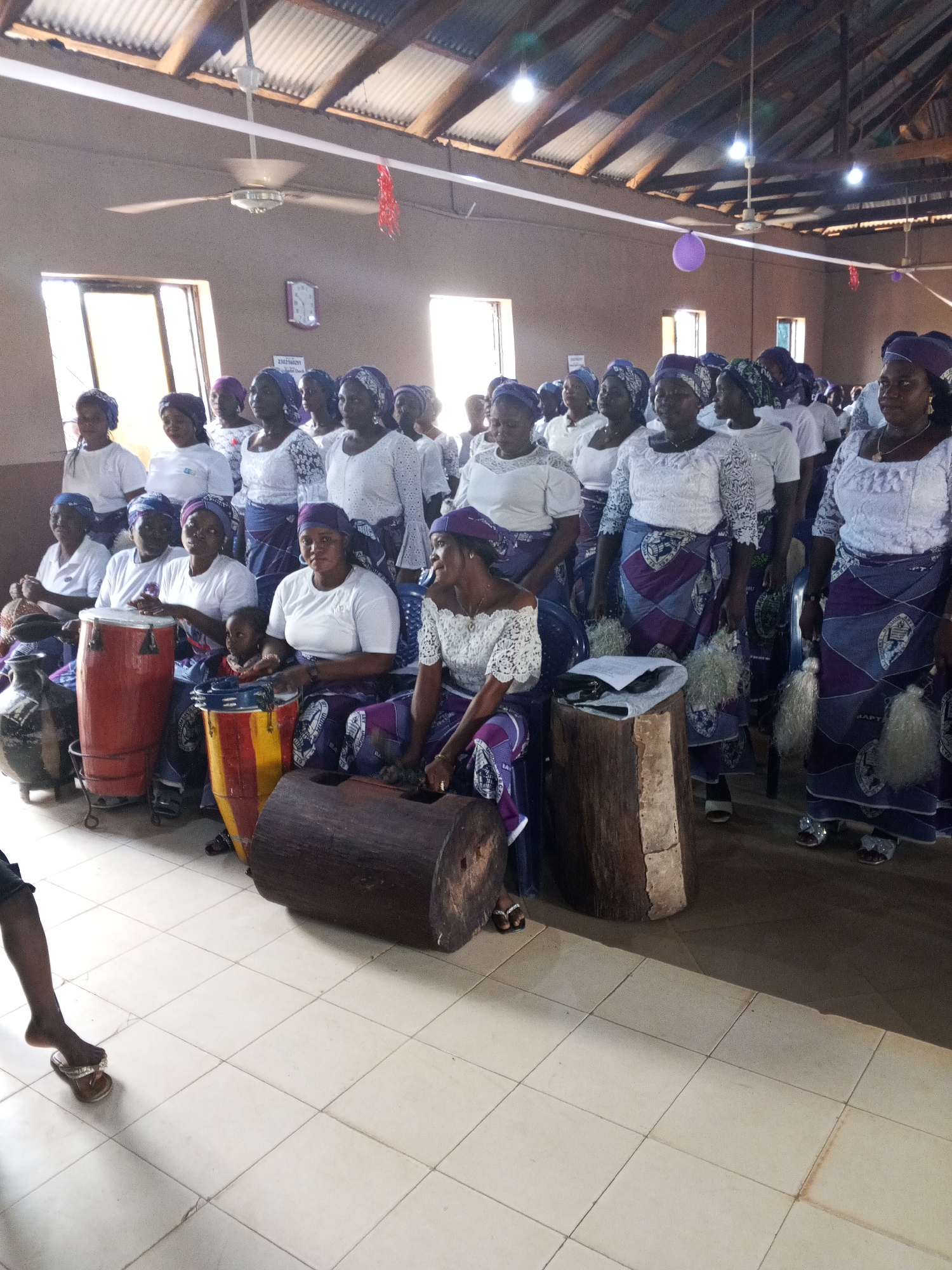 A group of approximately 25 women, likely a church choir, stand and sit in an indoor space, possibly a church in the city of Unknown, Unknown. The women are dressed in similar attire, consisting of white tops and purple and white patterned skirts, with matching headwraps. Some are playing drums, while others hold bundles of white fibers. The room has a corrugated metal roof, tile floors, and windows providing natural light. Overhead, ceiling fans are visible. Decorations, including a purple balloon and red streamers, adorn the space. A sign is affixed to the wall. A foot wearing a sandal is visible in the foreground.