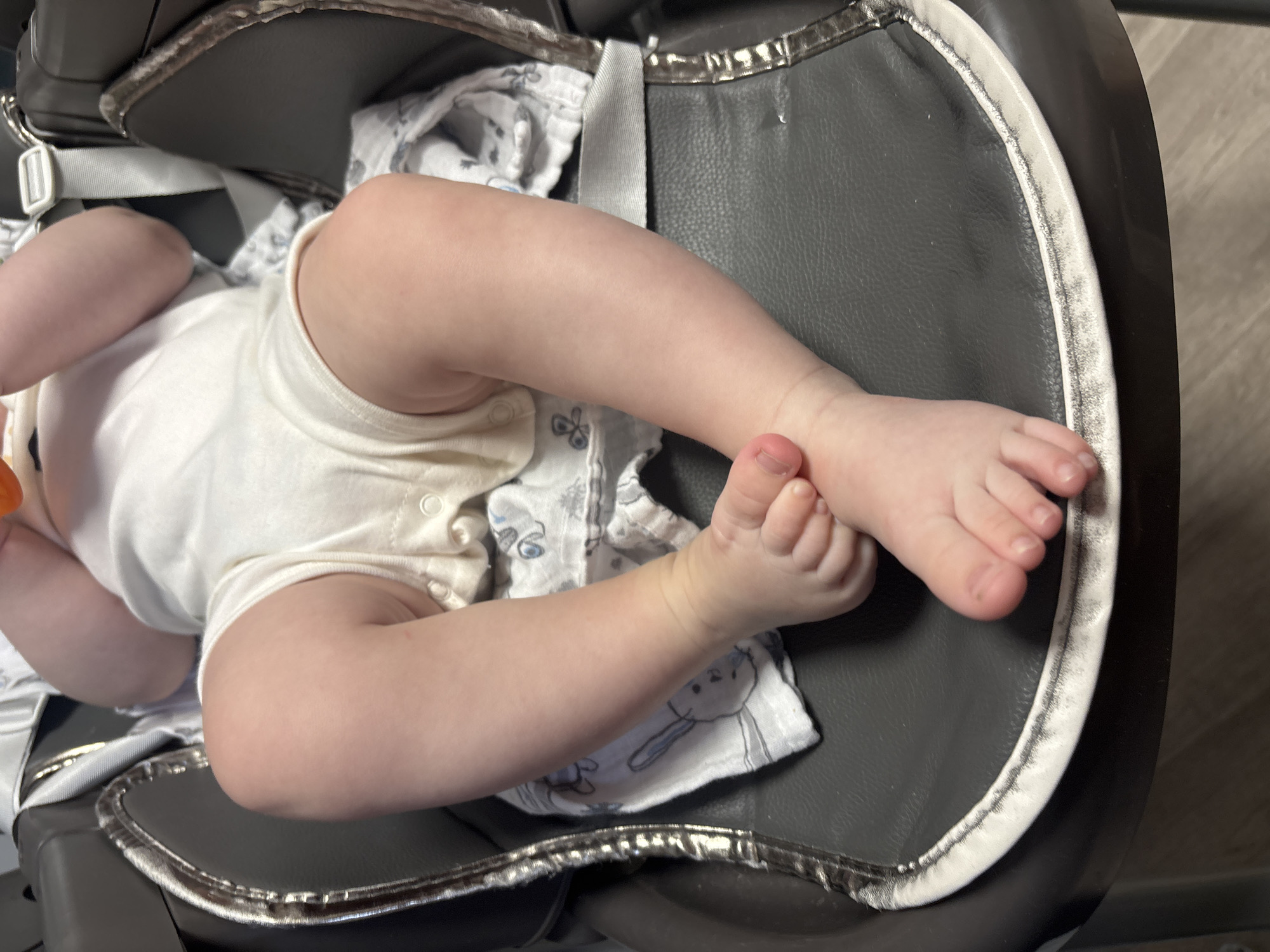 The image shows a baby lying in a gray high chair. The baby is wearing a white onesie with a patterned swaddle cloth. The high chair has a silver trim. The background appears to be a room with wood flooring. The location is unknown.