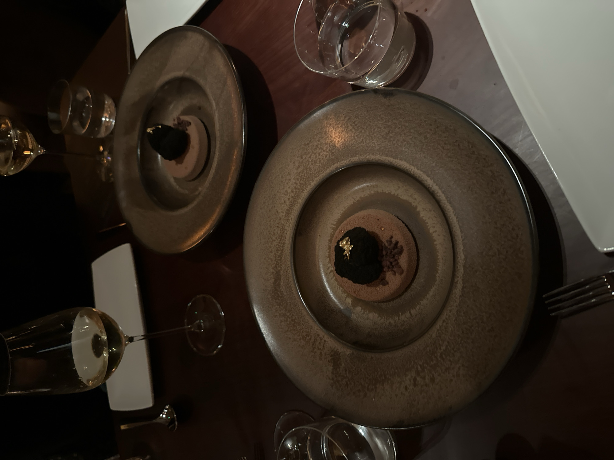 The image depicts a table setting, likely in a fine dining establishment in London, United Kingdom. Two ornate plates are visible, each containing a dessert. The dessert consists of a brownish base topped with a dark, irregular mound, possibly chocolate or mousse, adorned with small flecks of gold leaf and dark sprinkles. One plate is positioned closer to the viewer, revealing the details of the dessert more clearly, while the other is further away and partially obscured. Clear glassware, including water glasses and wine glasses filled with a light-colored liquid (presumably wine), are scattered around the plates. A white rectangular plate and a fork are also present, suggesting a multi-course meal. The table itself has a dark, polished surface, possibly wood. The lighting is dim and focused on the food, creating a sophisticated and intimate atmosphere.