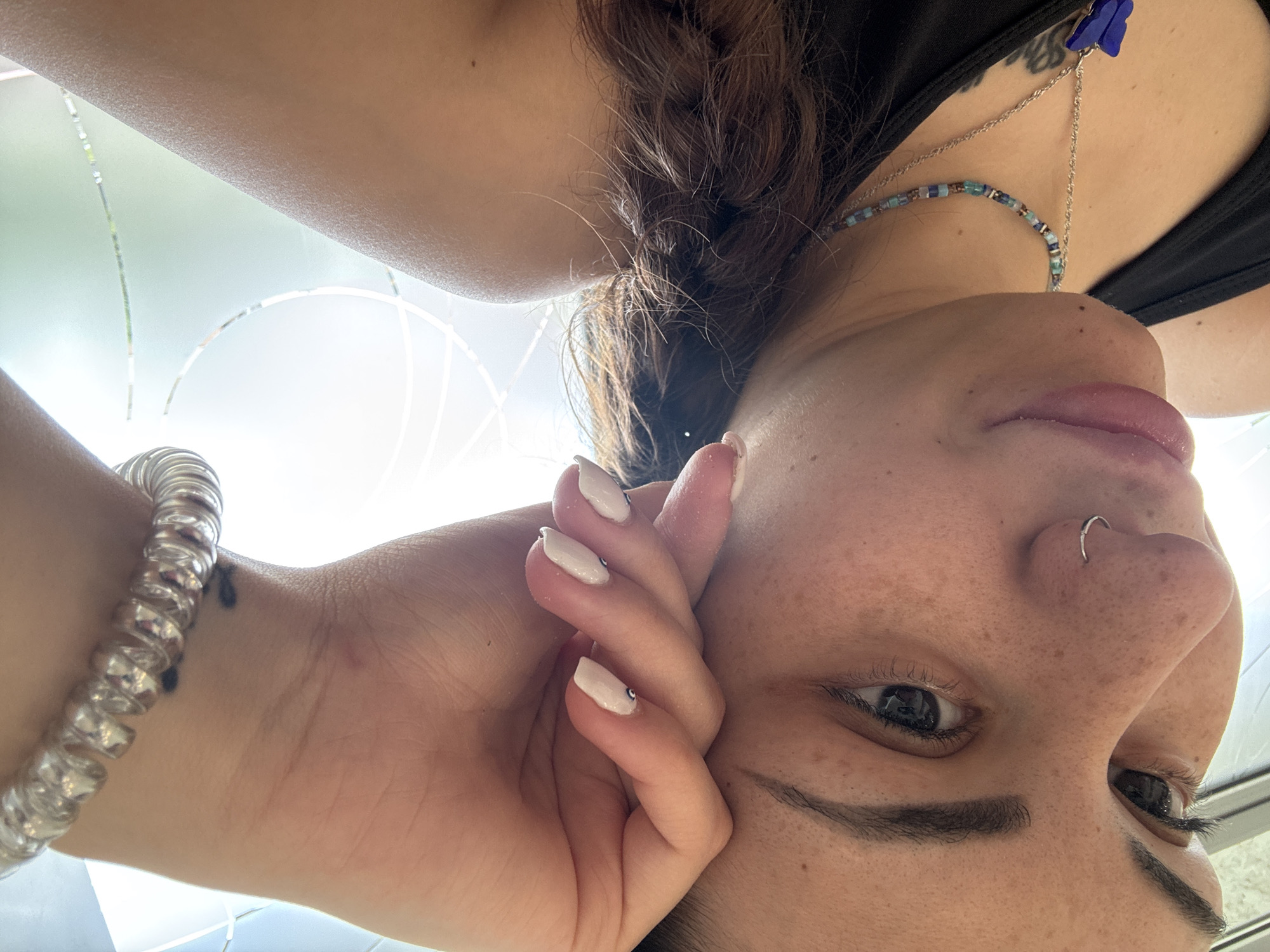 A close-up, inverted, worm's-eye view captures a person, likely female, looking downwards into the camera lens. The subject's face dominates the frame, showcasing freckled skin, dark eyebrows, and dark eyes. A silver hoop nose ring is visible on her right nostril. Her lips are slightly parted, and her dark brown hair appears tied back or braided.
She wears a black top, with a beaded necklace featuring blue, brown, and beige beads around her neck. A silver chain with a blue butterfly-shaped clip or charm is also visible near her shoulder. On her left wrist, she wears a clear or silver spiral-shaped bracelet, with a small dark tattoo faintly visible underneath. Her left hand rests near her face, revealing light-colored, manicured nails, some adorned with small, sparkling embellishments.
The background is brightly lit, suggesting natural daylight, and consists of soft white and pale green tones with faint, curved, and straight lines, possibly from a patterned window, frosted glass, or architectural features. The setting appears to be an indoor or sheltered outdoor space in Sofia, Bulgaria. The overall mood is casual and relaxed. There is no visible text in the image.