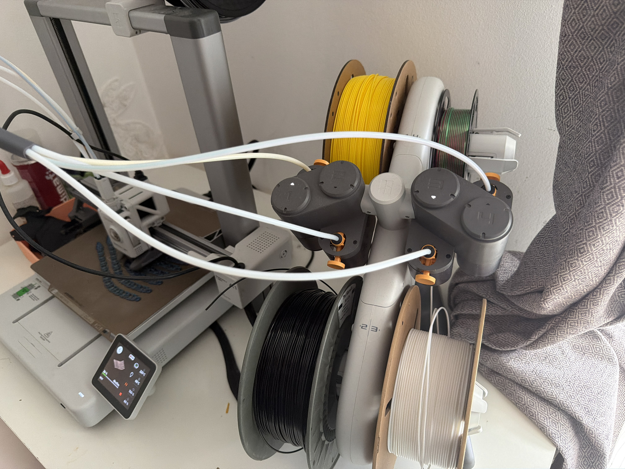 This detailed image captures a 3D printing setup situated on a white tabletop in the city of Unknown, Unknown. At the center is a light gray 3D printer, with a brown flexible build plate hosting a partially fabricated blue object. A small, square digital display, angled on the printer's base, shows real-time print parameters: 24% completion, 2047m filament used, 220°C nozzle temperature, 60°C bed temperature, 100% fan speed, and 60% flow rate. To the right, a gray multi-material system (AMS) houses four spools of filament: yellow (top, with wooden sides), iridescent green/red (middle, with white plastic sides), black (bottom-left, with gray plastic sides), and white/cream (bottom-right, with wooden sides). White Bowden tubes connect this system to the printer's print head. On the far left, a red can labeled 'dimati' and a white bottle with a red cap are visible, alongside black cabling. A patterned, diamond-weave fabric, possibly a curtain or blanket, drapes down the right side of the frame, contrasting with the white wall in the background.