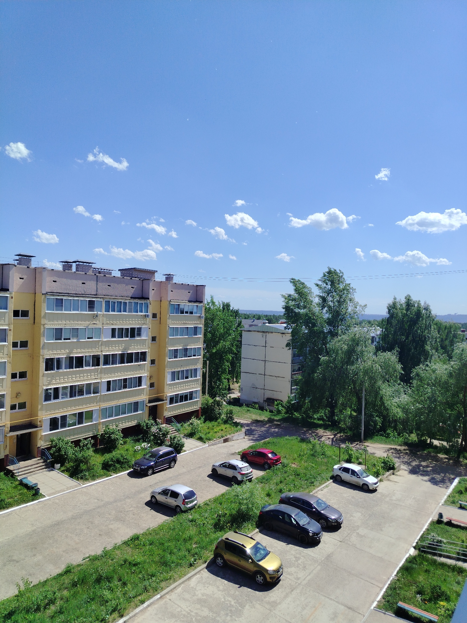 The image depicts a sunny daytime scene in what appears to be a residential area in Noviy, Russia. The sky is a clear blue with scattered white clouds. Dominating the left side of the frame is a tall, multi-story apartment building with a yellow and beige exterior. It has rows of identical windows and multiple chimneys on the roof.
In the foreground, there is a concrete parking area with several parked cars of various makes, models, and colors: gold, black, gray, red, white, and dark blue. The parking area is surrounded by grassy areas with patches of overgrown grass and weeds, indicating a lack of recent maintenance.
Behind the apartment building and parking area are more trees, another apartment building, and glimpses of the distant cityscape in the background. The overall impression is of a typical urban landscape in a Russian city. The image doesn't include any people or actions. The scene is static and focuses on the architectural and environmental elements.