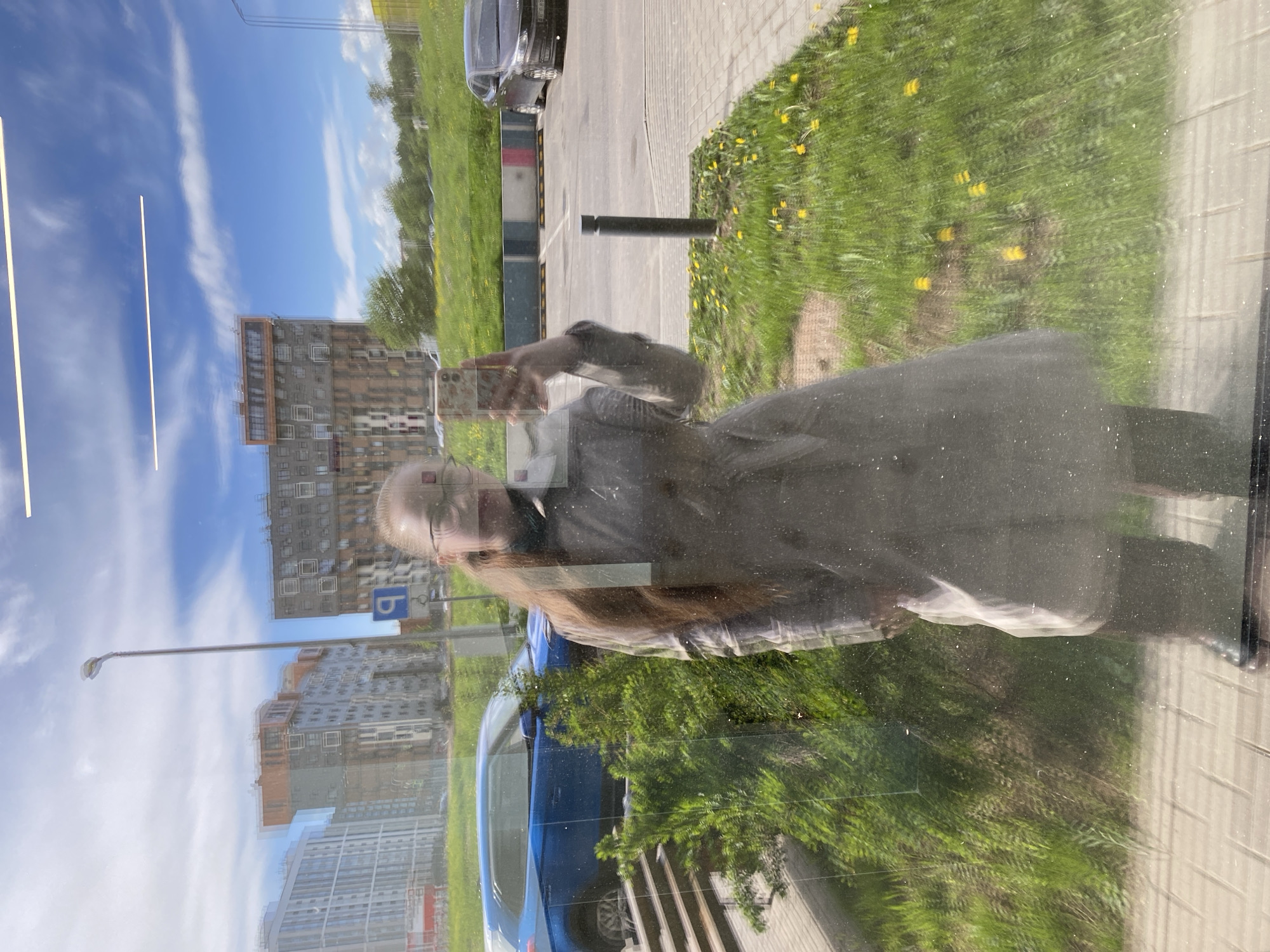 The image shows a reflection in a window. The reflection includes a person holding a smartphone, possibly taking a picture of themselves. In the background of the reflection are buildings, a blue car, grass, and the sky. Based on the buildings and overall environment, the location appears to be in a city, possibly Saint Petersburg, Russia. The reflection on the window obscures finer details. The presence of the person with the smartphone suggests an interaction with the environment through photography.