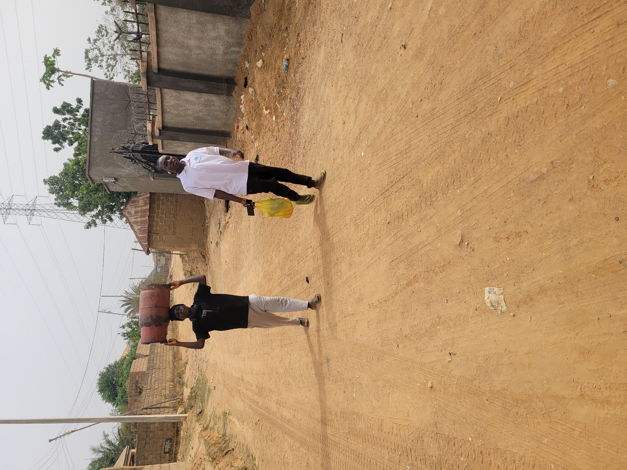 This image captures a daytime scene in Barakin Akawo, Nigeria, featuring two men walking along a dusty, unpaved dirt path.
On the left side of the path, a man is shown walking while expertly balancing two reddish-brown, possibly metal, barrels stacked on his head. He uses his hands to steady them. He is dressed in a black t-shirt with partially visible text (possibly "T.L."), light-colored trousers, and flip-flops.
Further along the path, to the right, a second man is visible, also walking. He wears a white short-sleeved shirt with a circular logo on the left chest area, which appears to contain the words "SILVER" or "SHERIFF" and "GLOBAL". He has black trousers and flip-flops, and carries a bright yellow plastic bag in his left hand.
The setting appears to be a semi-urban or rural area. The path itself shows tire tracks and is strewn with small debris. The background is dotted with various buildings, some constructed from unplastered concrete blocks, and others with grey plastered walls. One building features barbed wire along its top edge. Utility poles, electrical lines, and a large metal transmission tower are prominent features against the clear, bright sky. Green foliage from trees and bushes, including a distinct papaya tree, adds some color to the dry landscape. On the ground, near the man in the white shirt, a piece of crumpled paper is visible with the text "OUR NATION" partially legible. Another small sign on a building wall appears to have "HOTEL" written on it.
The strong daylight suggests it is mid-morning or afternoon, and the environment appears dry and sunny. The scene depicts everyday activities in the local environment.