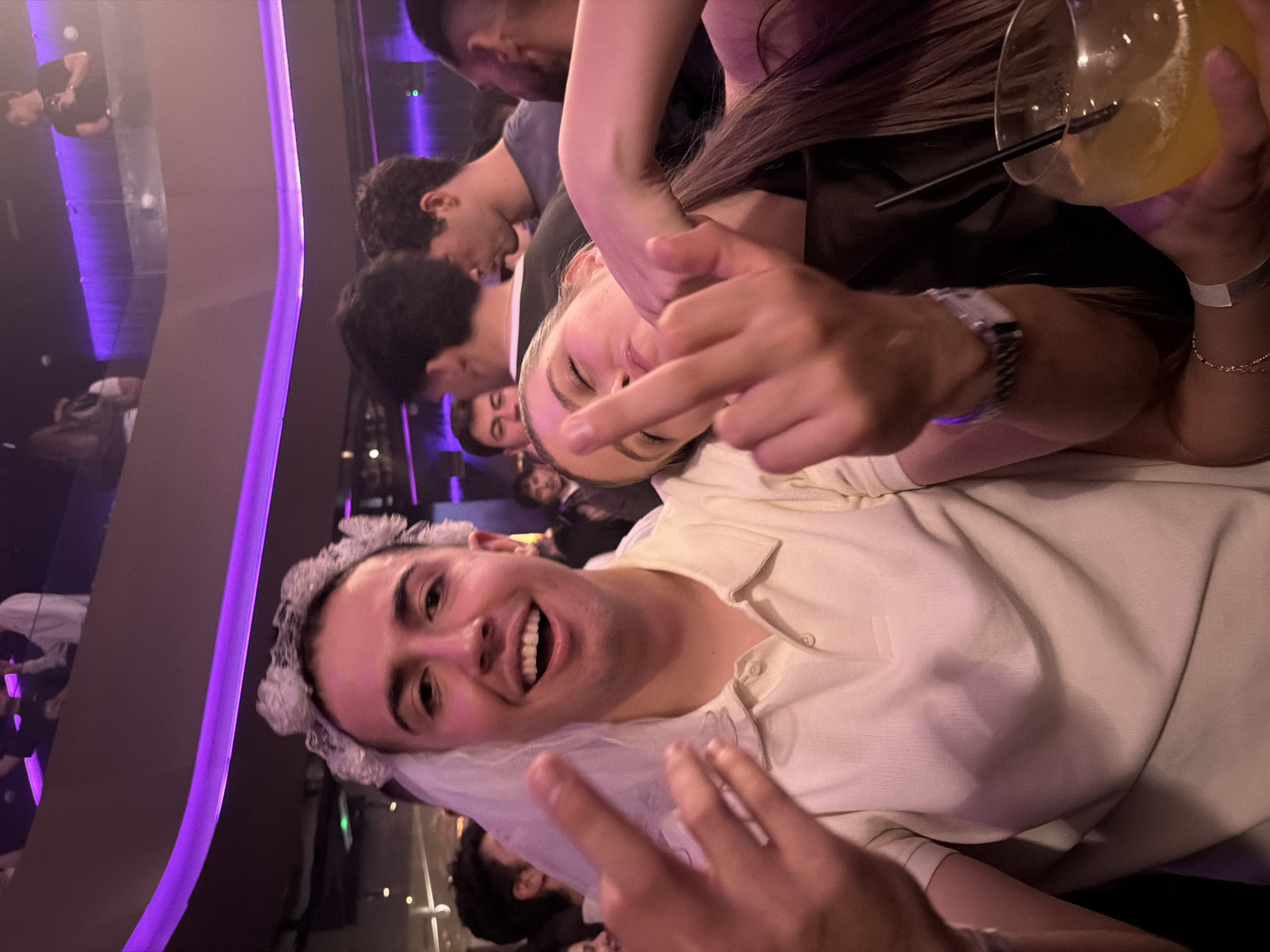 The image depicts a group of young adults inside a dimly lit nightclub, potentially located in the coastal city of Dénia, Spain. The focal point is a smiling male wearing a bridal veil and a light-colored polo shirt, gesturing with a peace sign. Nearby, a woman is shown giving the middle finger, while holding a cocktail glass with an orange liquid. The nightclub features a modern design, with curved walls illuminated by vibrant purple lighting. Various other individuals can be seen in the background.