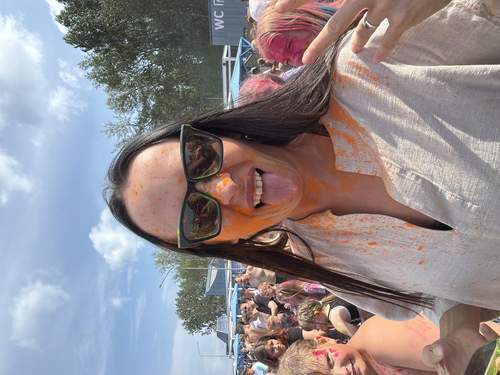 This is a medium shot photograph taken outdoors at eye level, featuring a woman with orange powder on her face and shirt. She is wearing large, dark-framed sunglasses and sticking her tongue out. Her long, dark hair is visible, as are parts of her shoulders and chest. The background shows a crowd of people, some of whom also have colored powder on them. Trees and a blue and white tent structure are visible in the distance, along with a grey shipping container with "WC" printed on it. The sky is blue with scattered white clouds. The event appears to be a color festival, likely taking place in Chaikovsky, Russia, given the context. The woman's right hand is raised, and she is making a peace sign with two fingers.