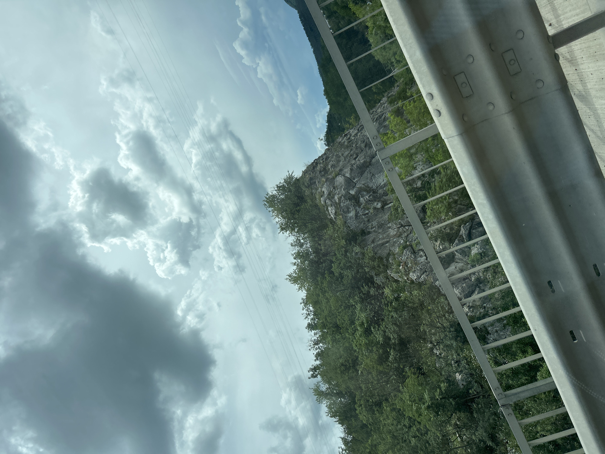 The media file displays a scene from a moving vehicle, likely a car, looking out towards a mountainous landscape. The image is rotated, with the sky occupying the left side and the terrain on the right. A prominent feature is a silver guardrail running along the right edge of the frame, appearing to be part of a road or bridge.
The sky is filled with dramatic, cloudy formations, with some areas of bright sunlight breaking through, suggesting a partly cloudy or overcast day. A series of thin power lines are visible stretching diagonally across the sky, adding a man-made element to the natural scenery.
The terrain consists of steep, rocky slopes covered in dense green foliage, indicative of a forested mountainous region. The rocks are grey and jagged, interspersed with trees and smaller shrubs. The vegetation appears lush and healthy, suggesting a temperate climate.
The overall impression is one of travel through a scenic, natural environment. The time of day is difficult to pinpoint precisely but the lighting suggests daytime. There are no people visible in the image. No text is present.