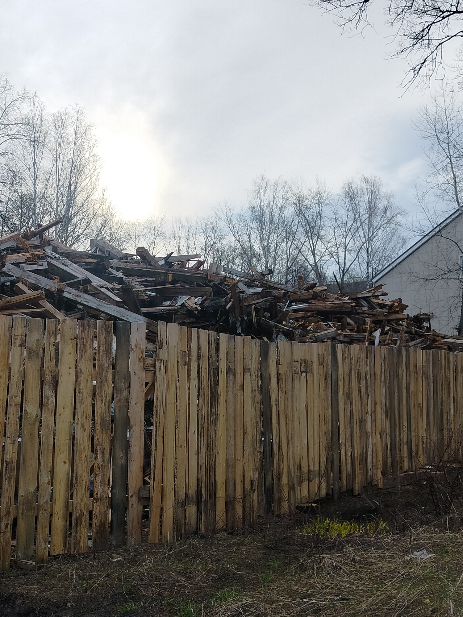 The image depicts a large pile of what appears to be discarded wood debris stacked behind a wooden fence. The location is likely near the city of Chaikovsky, Russia.
The fence is constructed of vertical wooden planks. The wood debris behind the fence consists of various shapes and sizes. In the background, bare trees and a building with a slanted roof are visible against an overcast sky. The ground in front of the fence is covered with dead grass and sparse green vegetation.