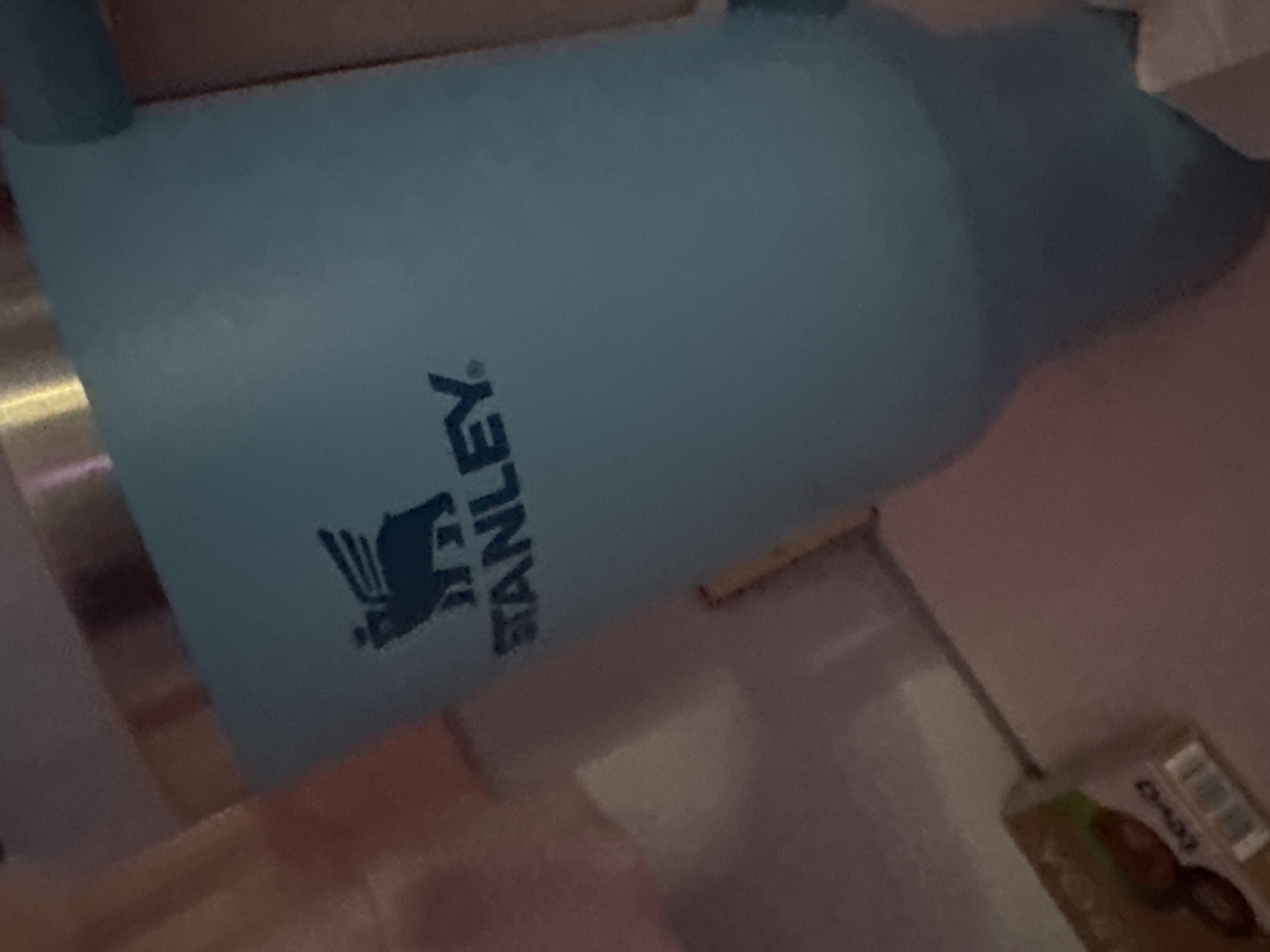A light blue Stanley brand insulated tumbler, featuring its distinct winged bear logo and "STANLEY" text in dark blue, occupies the majority of the frame. A metallic band is visible at the top of the tumbler. The background is an out-of-focus indoor environment with warm, dim lighting. To the lower left, a brown cardboard box, partially labeled "ROA" and "teva" with a visible barcode, rests on a surface. No individuals or active interactions are present in the image. This scene is located in the city of Rezbartsi, Bulgaria.