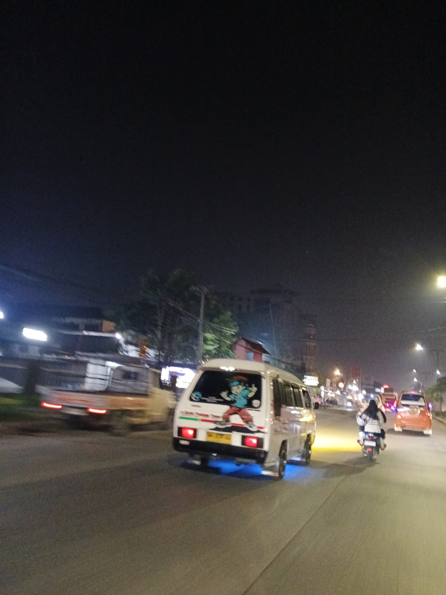 The image depicts a nighttime street scene in Medan, Indonesia, focusing on vehicular traffic. In the foreground, a white minivan with a cartoon decal on its rear side is moving on the right lane of a road. The minivan has blue underglow lighting. To the right of the minivan, a person riding a motorcycle is visible, also moving in the same direction. Further down the road, another vehicle, possibly an orange car, is seen in the distance. The road appears to be asphalt, and streetlights are illuminating the scene. In the background, buildings and trees are faintly visible, shrouded in the darkness of the night. The overall impression is of a busy urban street at night, with vehicles navigating the road.