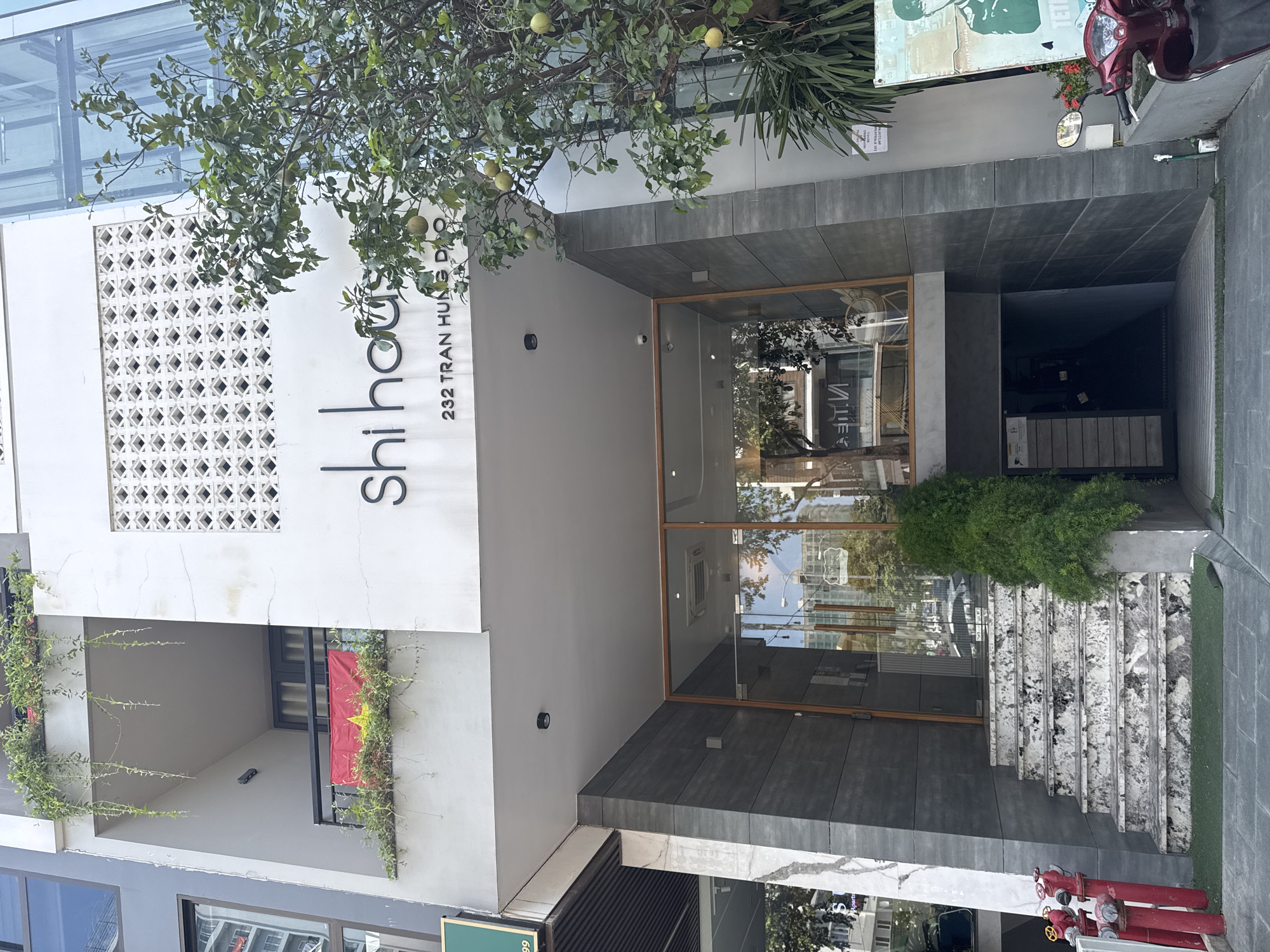 The image shows the facade of "Shihouis" cafe located at 232 Tran Hung Dao Street in Đà Nẵng, Vietnam. The building is characterized by a modern design with a white upper facade featuring a geometric lattice pattern and the cafe's name and address in black lettering. A tree with green fruit overhangs the entrance area. Below this section is a window with the Vietnamese flag. The entrance itself is a glass-fronted doorway set back from the street, with a small manicured bush and marble steps leading up to it. In front of the steps, there is the top of a fire hydrant. A red scooter and a green sign are to the right of the building.