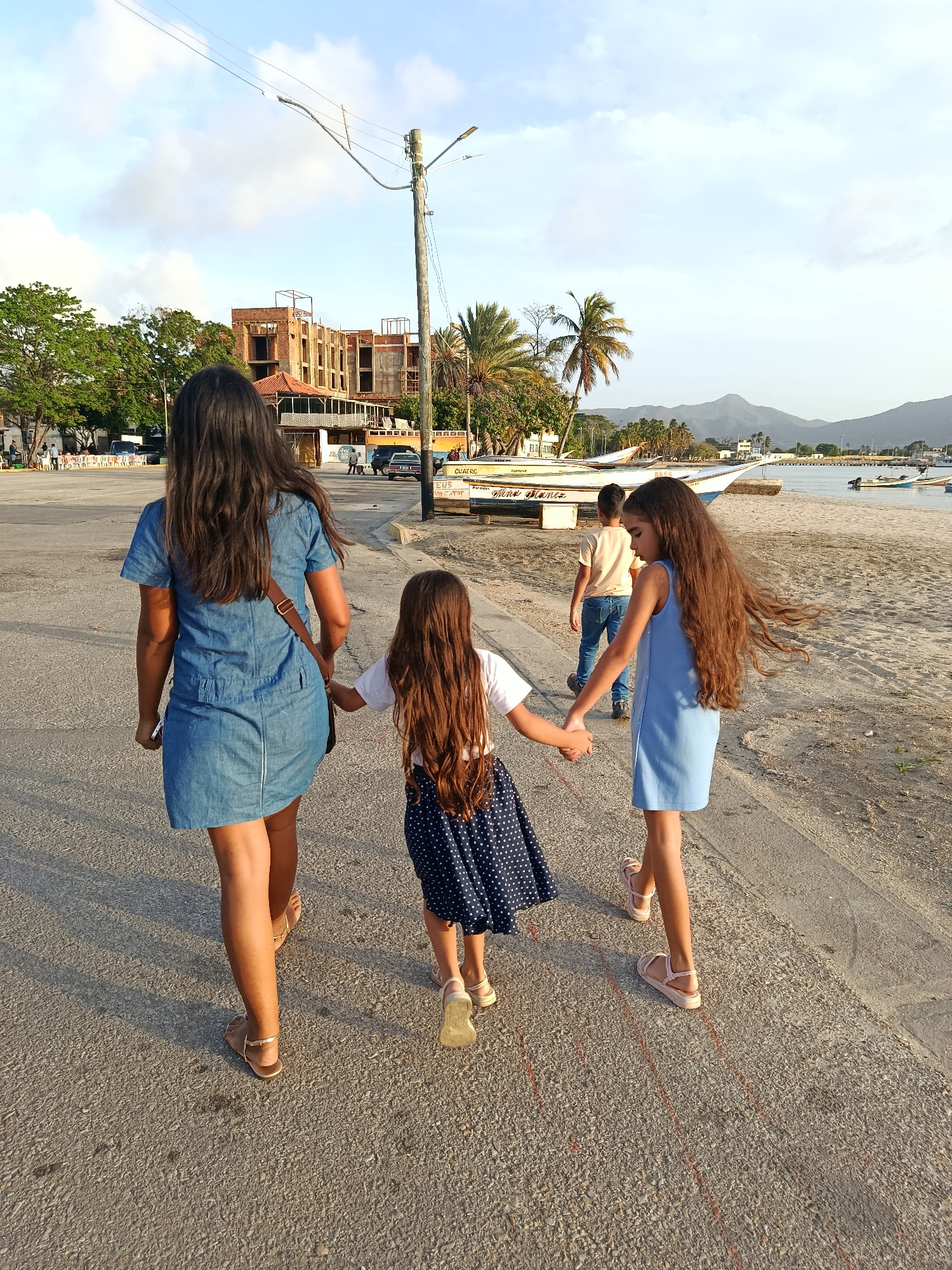 In Juan Griego, Venezuela, a group of four people, two adults and two children, walk along a paved path bordering a sandy beach. The adult on the left, a woman with long dark hair, wears a denim dress and a brown crossbody bag. She holds hands with a young girl in the center, who has long, dark, wavy hair and wears a white t-shirt and a polka-dot skirt. Beside her, another girl, also with long, dark, wavy hair, is dressed in a light blue dress and holds the hand of the central child. Behind them, a young boy in a light yellow shirt and blue jeans walks on the paved path. The background reveals a coastal town with a partially constructed building, palm trees, a street with parked cars and tables, and several boats docked in the water. Mountains are visible in the distance under a cloudy sky, suggesting late afternoon or early evening light.