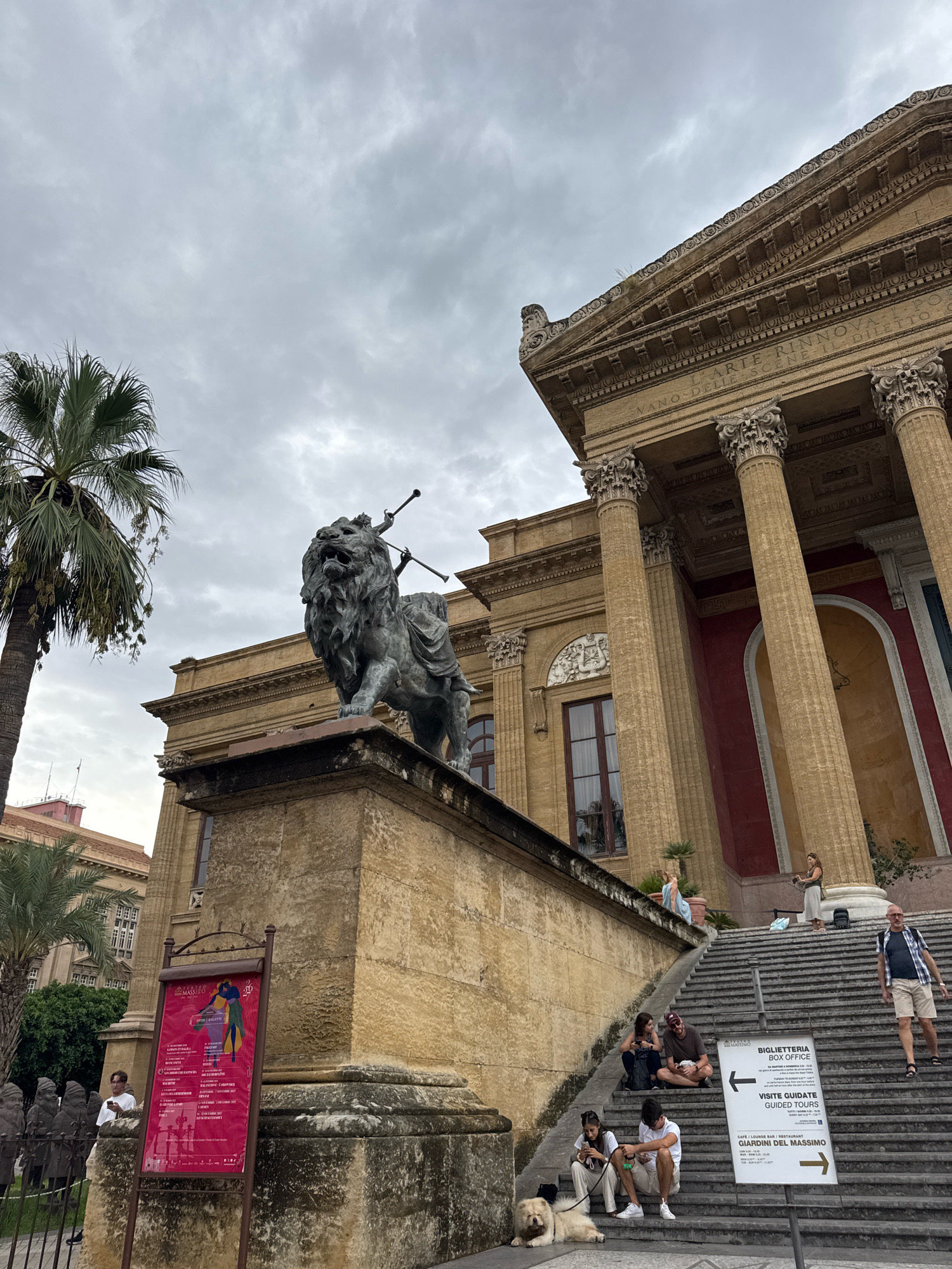 The image depicts a scene in front of the Teatro Massimo in Palermo, Italy, on an overcast day. A large, dark bronze sculpture of a lion, seemingly adorned with trumpets or horns, stands prominently on a stone pedestal to the left of the frame. The lion sculpture is positioned in front of the imposing classical architecture of the theater, characterized by large columns and a weathered stone facade.
A wide set of stone stairs leads up to the theater's entrance. Several people are visible on these stairs. In the foreground, a light-colored dog lies on the lower steps, next to a seated young woman holding its leash. Further up the stairs, a group of individuals are seated, some talking and others looking around. A man in shorts and a checkered shirt is walking down the stairs on the right side of the frame. Two women are standing on a higher ledge to the right, one appearing to take a photo.
A red poster advertising events is displayed on a stand to the left, near the base of the lion sculpture. A sign with directional arrows for "BIGLIETTERIA BOX OFFICE" and "VISITE GUIDATE GUIDED TOURS" is placed on the stairs, pointing towards the theater. Palm trees frame the left side of the image, with one tall palm tree reaching towards the cloudy sky. The overall impression is a lively public space outside a significant cultural landmark, with a mix of tourists and locals enjoying the area.