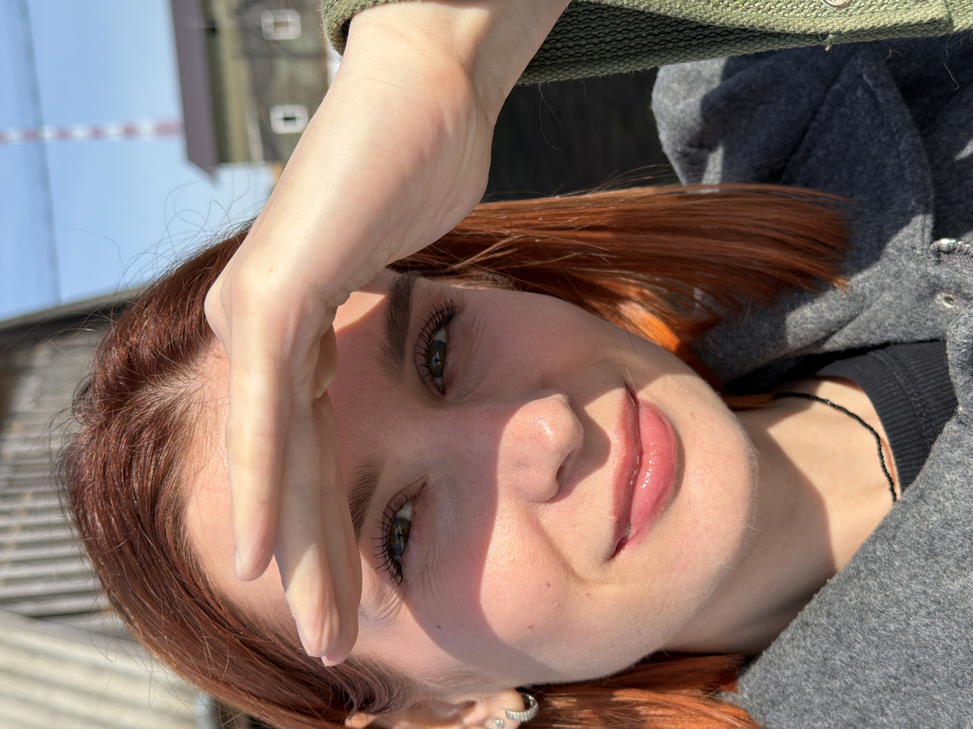 A young woman with red hair is facing the camera with her hand shielding her forehead from the sunlight. She is wearing a grey jacket and a black necklace is visible. The background includes buildings, trees, and a blue sky. The location is likely in Chaikovsky, Russia.
