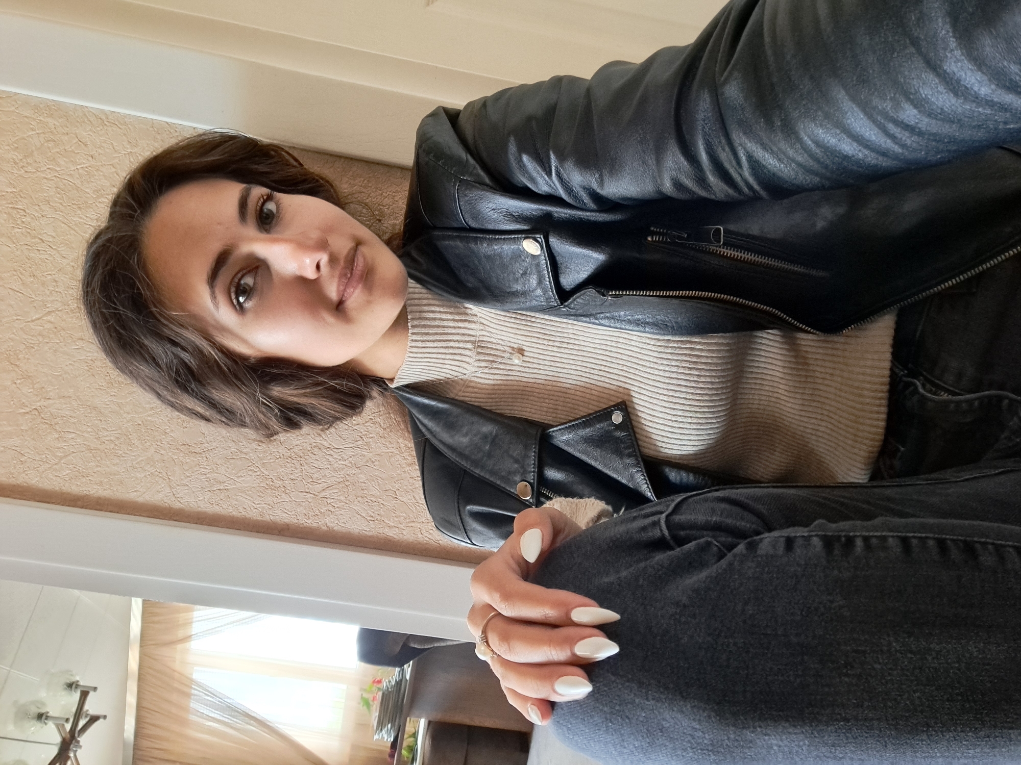 A woman poses indoors, wearing a black leather jacket over a tan ribbed top and dark jeans. Her dark hair is styled in short waves, and her fingernails are painted white. She is seated, with one hand resting on her knee. The background includes a beige wall and a glimpse of an interior space with a window covered by a translucent curtain. The city and specific location are unknown.