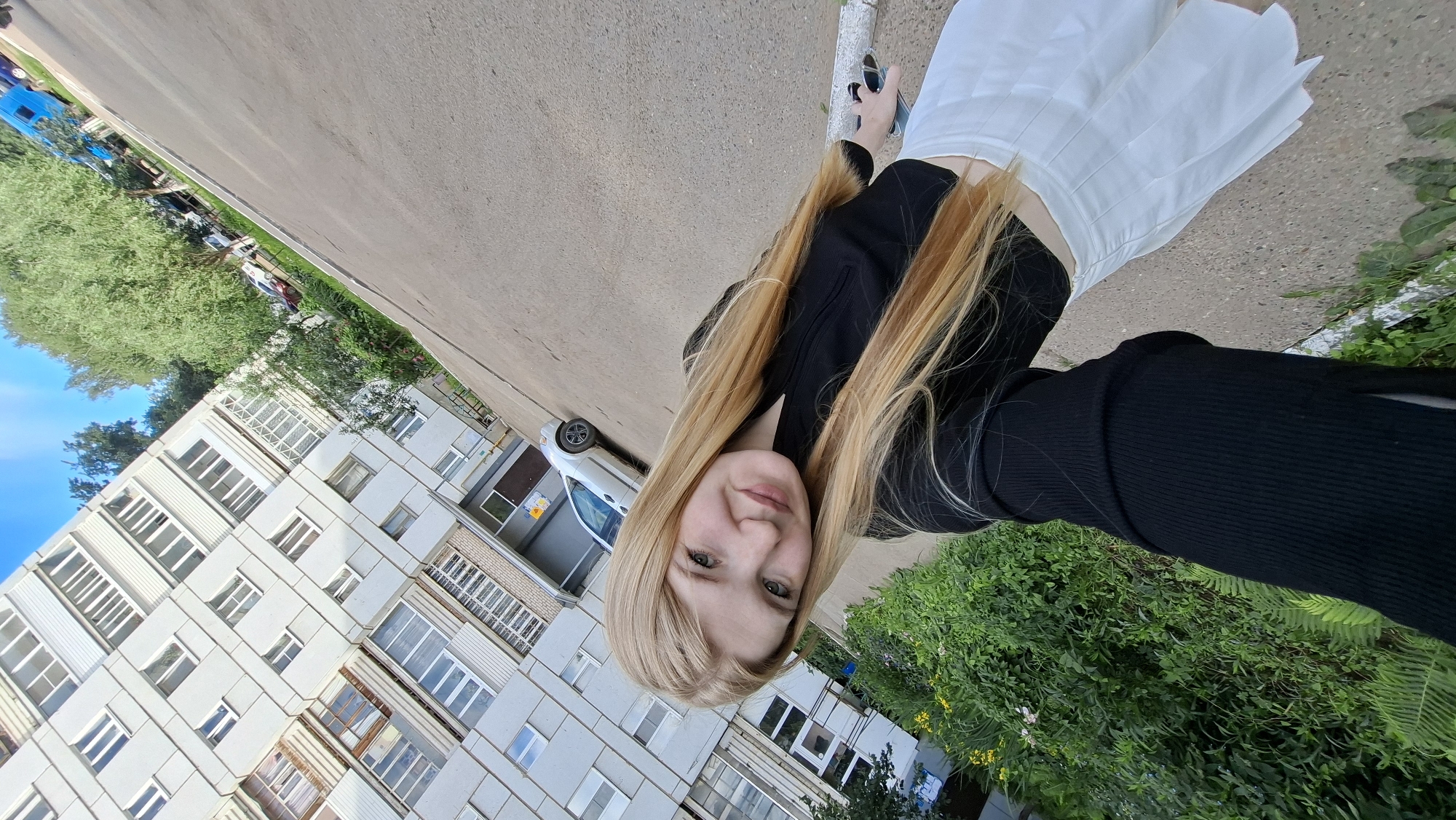 A young blonde woman stands on a sidewalk in front of a Soviet-era apartment building, likely in Chaikovsky, Russia. She is wearing a black long-sleeved crop top and a white pleated skirt. The building has a grey facade with multiple windows and balconies. To the left of the building, there is lush green foliage. Behind her, a silver car is parked on the pavement. The sky is blue with some clouds, and trees are visible in the background. The woman is holding a phone in her right hand, taking a selfie. The image is taken from a slightly tilted perspective.