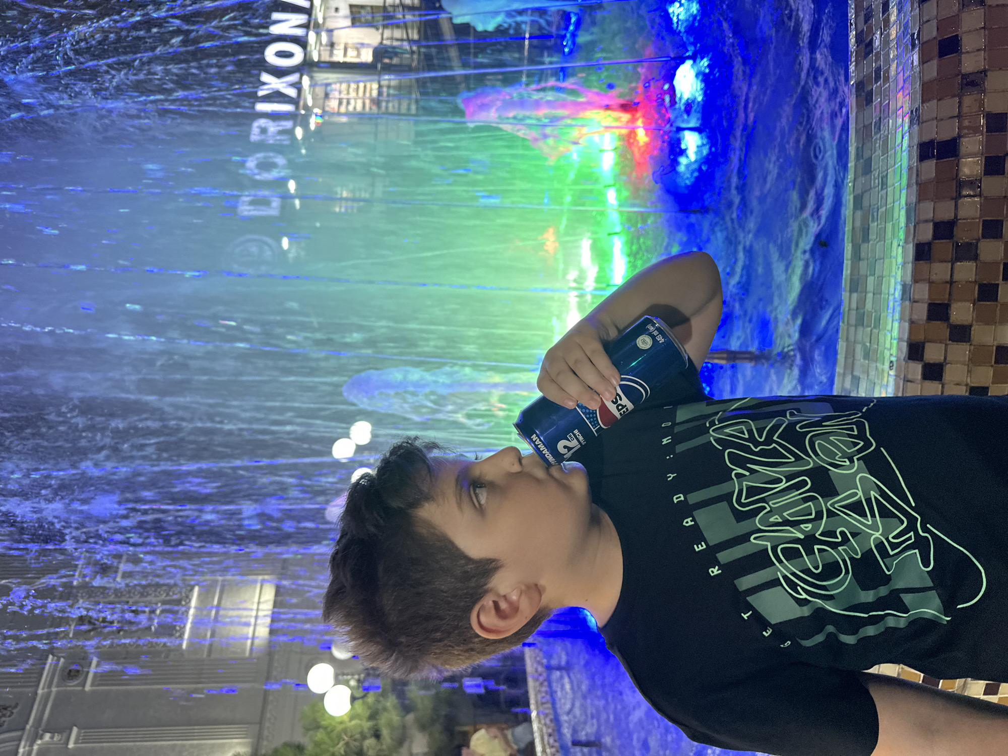 In Tashkent, Uzbekistan, a young boy stands in front of a vibrant, multi-colored fountain, holding a can of Pepsi to his lips. He is wearing a black t-shirt with the slogan "GET READY: GAME ON" emblazoned in a stylized, green font. The fountain's water is illuminated by blue, green, and pink lights, creating a dynamic and colorful backdrop. In the background, a building with the partial text "DORIXONA" is visible, indicating a pharmacy. The scene appears to be an outdoor evening setting, possibly a public square or entertainment area.