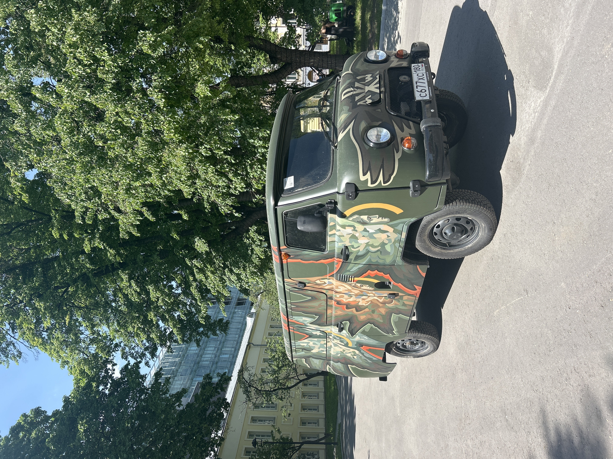 The image shows a green and brown camouflage-painted UAZ-452 van parked on a paved surface. The van has a Russian license plate "C677XC180" and is decorated with a large, colorful illustration that appears to be dragon-like, covering the side panel. A large green tree is visible in the background. Behind the tree, parts of buildings can be seen, including a modern glass-facade building and an older yellow building with windows. The location is in an unknown city, in an unknown region. The image seems to be taken during daytime, as indicated by the shadows cast by the van. The van appears to be the main subject in the scene.