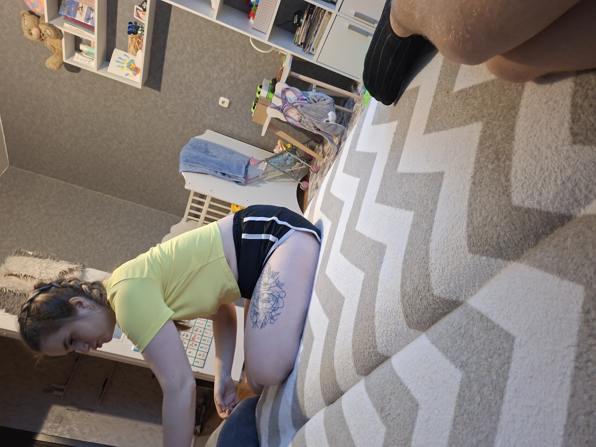 The image shows a room with shelves on the top-left, containing books and other items. Below the shelves, there's a wall. In front of the wall, there's a white highchair and a child's stroller. A person wearing a yellow shirt and black shorts with white trim, kneeling near a table. A tattoo of flowers is visible on their left leg. A sofa covered in a zig-zag patterned blanket dominates the right side of the image. A person wearing a sock is sitting on the sofa.