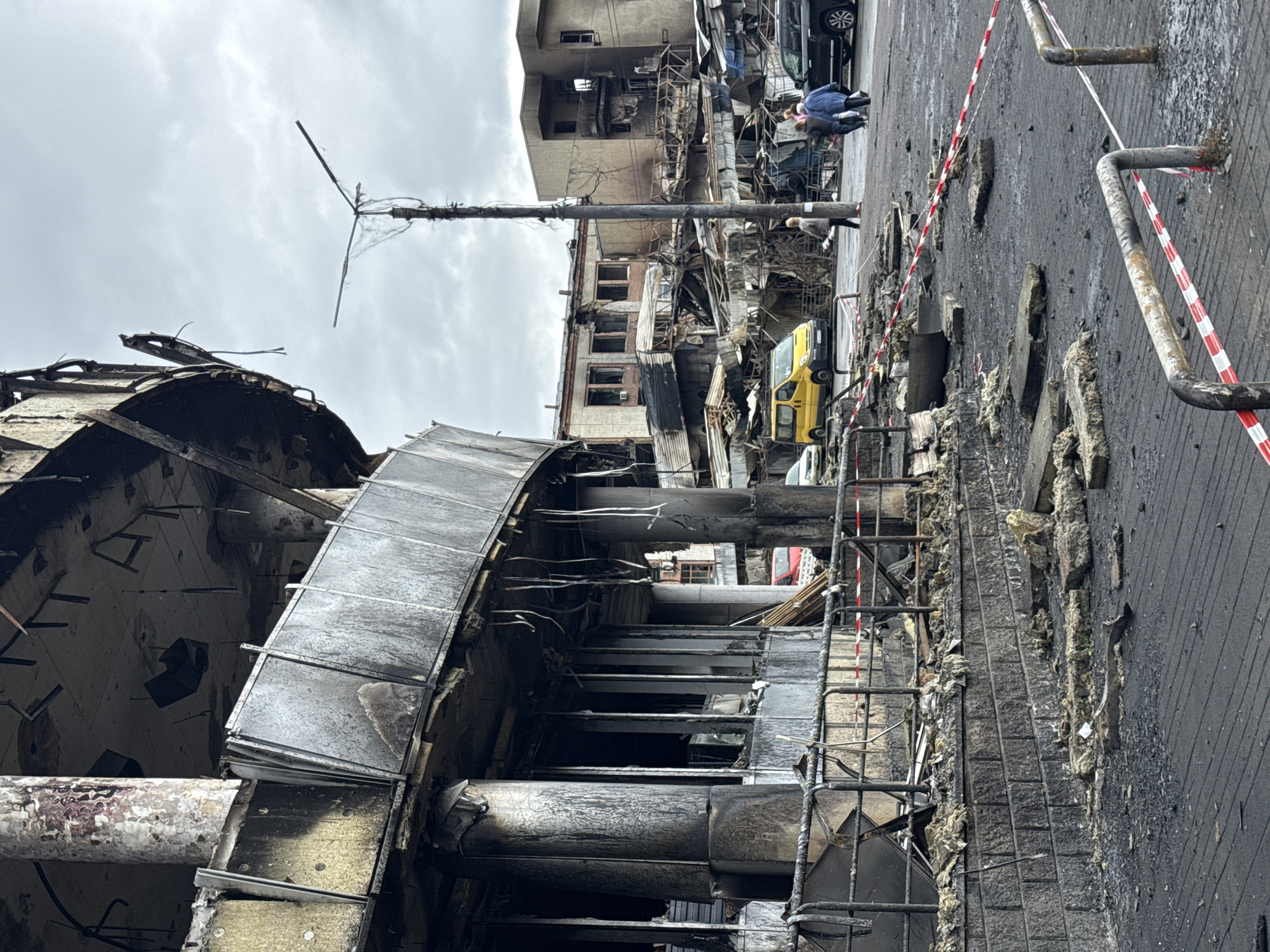 The image shows the aftermath of a blast in Kyiv, Ukraine. A severely damaged building with charred remains and exposed structural elements is prominent. Debris litters the foreground pavement, which is also cracked and burned. A red and white barricade tape is visible. In the background, another building is damaged, and a yellow van and other vehicles are parked nearby. Several people are visible in the background. Overhead, the sky is overcast. A damaged utility pole is also visible.