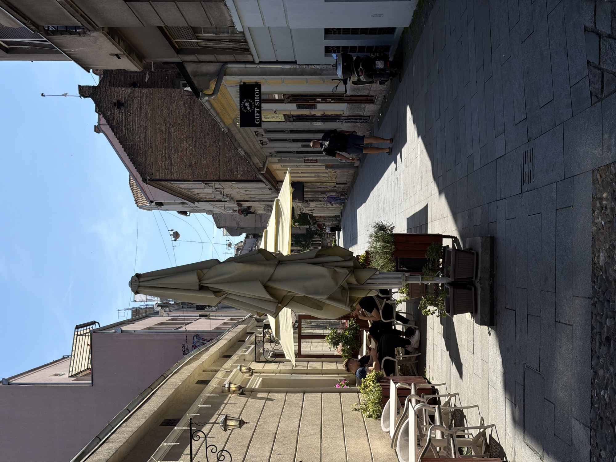 The image shows a street scene in the city of Beograd, Serbia, likely in a pedestrianized area or a narrow street with outdoor seating for cafes. In the foreground on the left, there are tables and chairs set up outside a building. Two young men are seated at one of the tables, facing each other, with one appearing to read. Several potted plants are placed around the seating area.
Further down the street, a large, folded umbrella is prominently positioned, casting a shadow on the cobblestone pavement. The street is lined with buildings on both sides, displaying a mix of architectural styles. On the right side of the street, a sign for a "GIFT SHOP" is visible above a storefront. A man wearing a black t-shirt and blue shorts is walking away from the camera down the street. A scooter is parked near the gift shop. The sky is clear and blue, suggesting a sunny day. The overall impression is of a lively street with commercial establishments and outdoor dining.