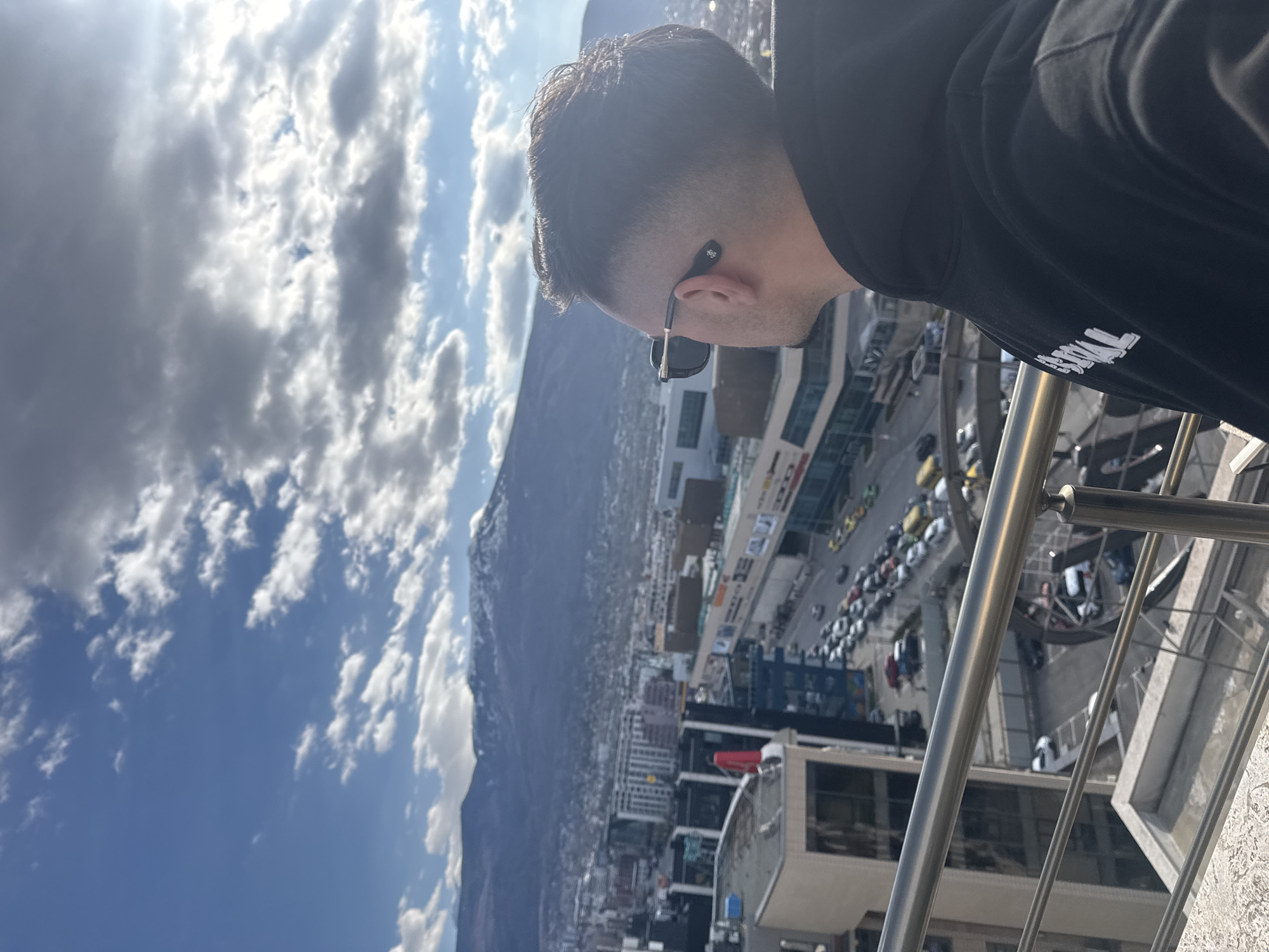 A man wearing a black hoodie and sunglasses stands on a balcony, looking down at the city of Sofia, Bulgaria. In the background, a large mountain rises above the cityscape. The sky is filled with clouds. Below, there are many buildings and streets.