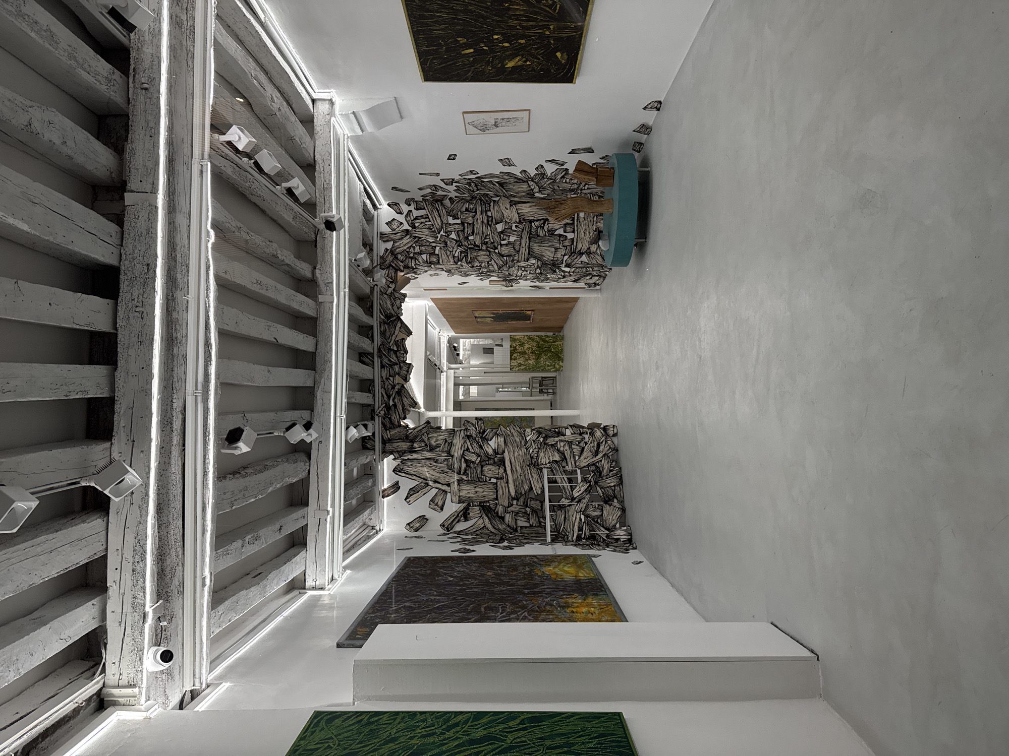 The image presents an interior view of an art gallery space, possibly located in Paris, France. The space features an installation made of what appears to be a collection of wood planks or drawings resembling planks that appear to be bursting from the walls, the ceiling and the floor. The gallery walls and floor are primarily white or light grey in color. On the left side, the wall has a wooden, beam-like structure with integrated lighting. Security cameras are mounted on this structure. Further down the hall is a ladder positioned against the wall. Artwork, possibly paintings, are mounted on the ceiling and floor. A curved sofa or seat is placed near the far end of the hall on the right side. The overall scene gives a minimalist and artistic impression. The space appears to be empty of people at the time the photo was taken.
