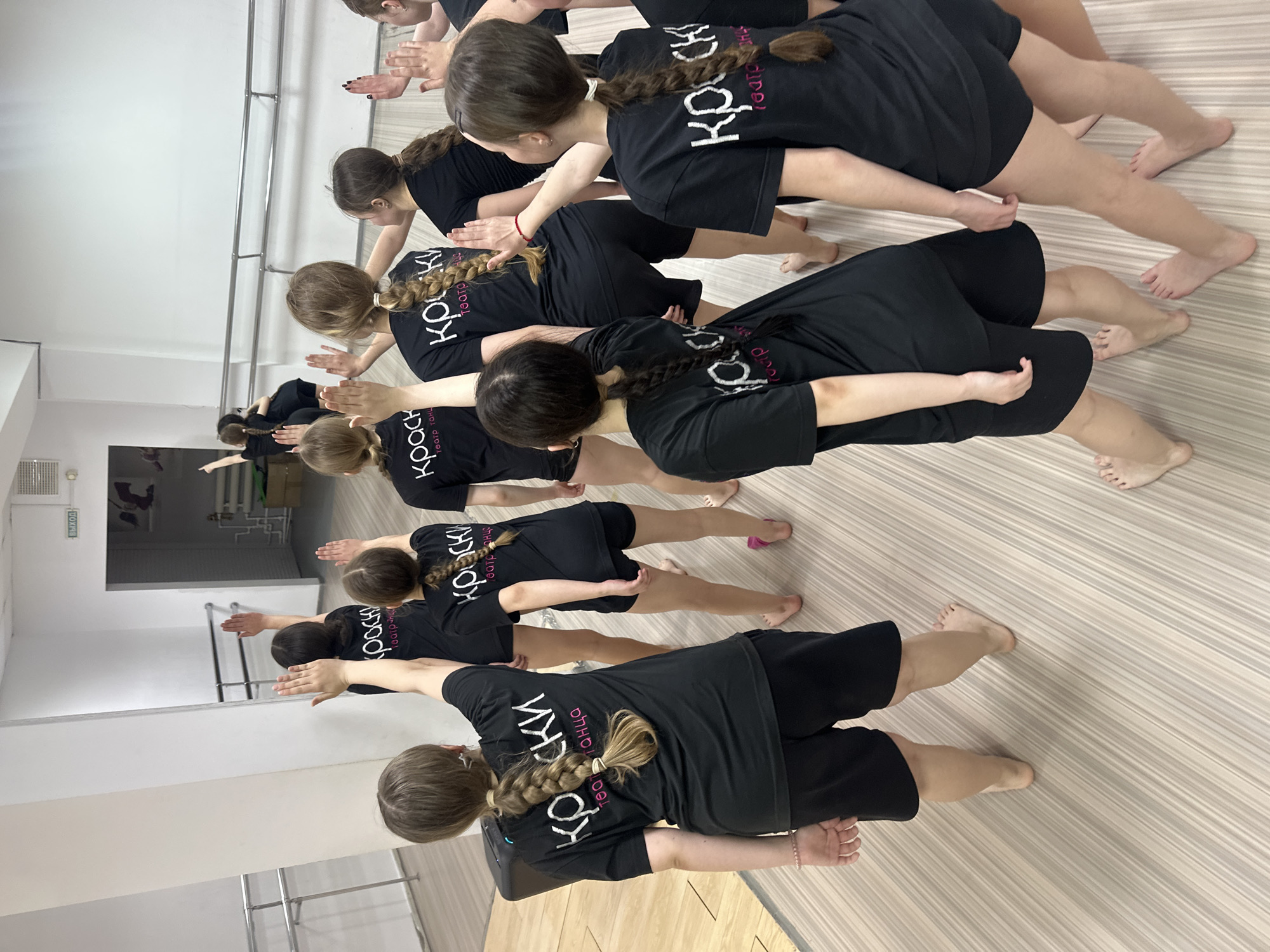A group of young people in black t-shirts are practicing a dance or exercise routine in a studio. The studio appears to be in Votkinsk, Russia based on the text on their shirts. They are lined up in rows and are barefoot. Their t-shirts have the name "КРОСКИ" with the text "театр танца" below it in white and pink letters. They are stretching with their arms extended. The studio has a mirror along one wall, a barre, and a light-colored wood-patterned floor. There is a door visible through the mirror.