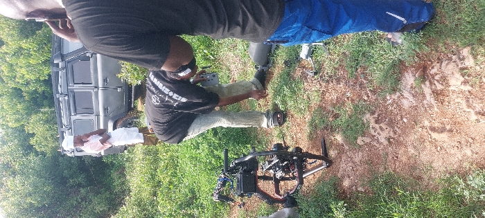 This image depicts an outdoor scene in Dar es-Salaam, Tanzania, characterized by unpaved earth, interspersed green grass, and dense foliage from trees and bushes. The setting appears to be a natural or undeveloped area.
Three adult males are visible. In the foreground right, a person is partially visible from the chest down, wearing a black t-shirt and blue cargo pants, with their right arm raised towards their head, appearing to hold a phone or adjust an earpiece. Directly in front of them, slightly to the left, another individual is bent at the waist, facing downwards. This person wears a black t-shirt with white graphics, light-colored pants, and black sandals, holding a grey remote control with both hands and looking towards a small grey drone resting on the ground near their feet. A white band is around their head or neck. To the far left in the midground, a professional camera rig on a black gimbal stabilizer is positioned on the ground. In the background left, another man stands upright, dressed in a light pink button-down shirt, white shorts or a similar garment, and dark footwear. He wears a white head covering, has his arms crossed over his chest, and looks towards the left side of the frame. Behind him, a dark grey Land Rover Defender SUV with a roof rack and a snorkel is parked amidst the vegetation. The scene suggests a film or drone operation in progress or preparation.