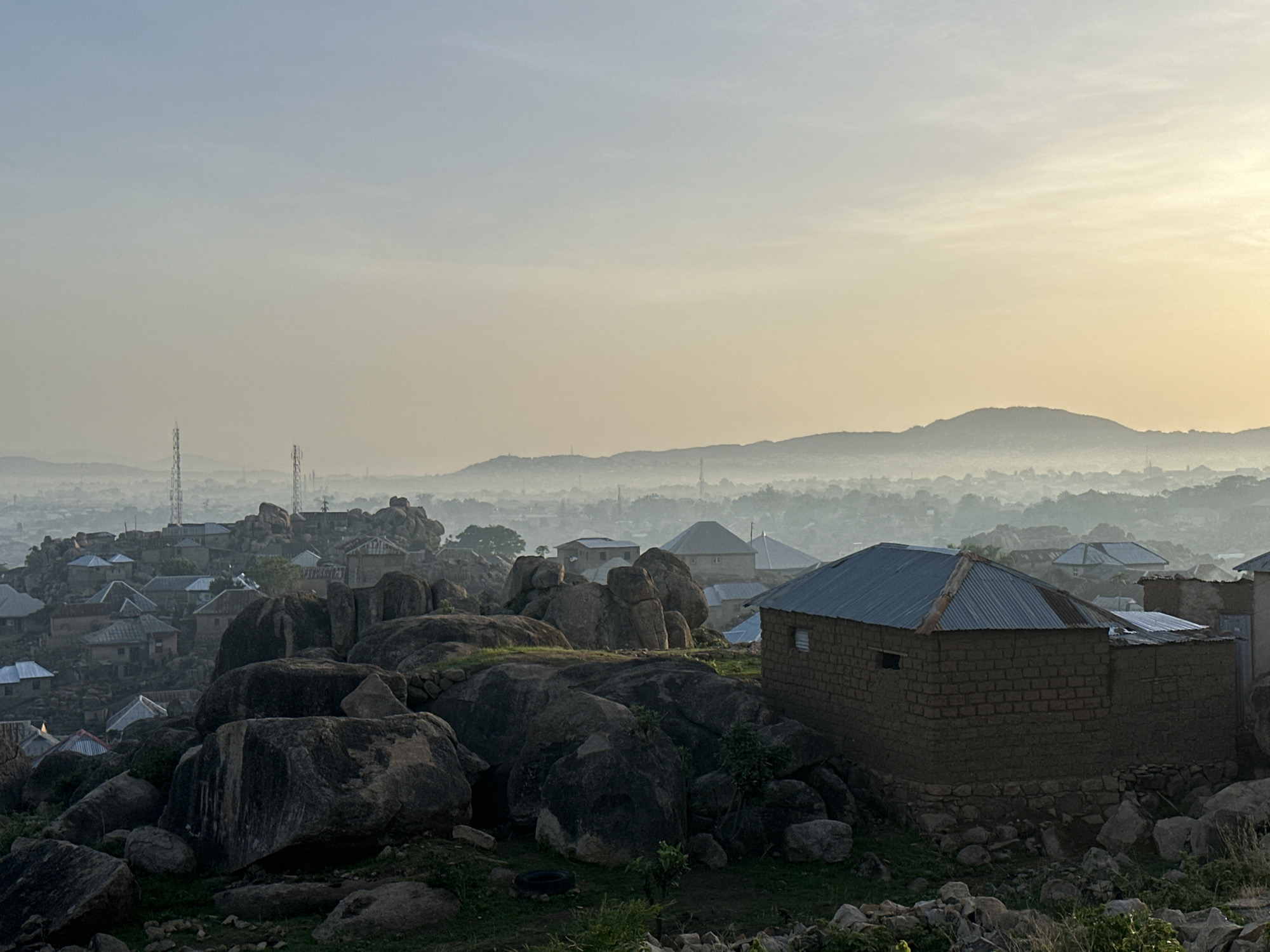 This panoramic image presents a wide view of a town in Jos, Nigeria, nestled within a distinctive rocky landscape under a hazy sky. The soft, warm light and the gradient of the sky, shifting from pale blue on the left to a golden-yellow glow on the right, suggest either early morning or late afternoon, likely sunrise or sunset.
In the foreground, large, dark, weathered boulders dominate the terrain, interspersed with sparse green vegetation. Scattered among these rocks are numerous single-story houses, constructed with mud brick or similar materials and topped with corrugated metal roofs, mostly silver or grey. A prominent house in the mid-right features a brown-bricked base, mud-brick walls, and a rusty corrugated iron roof.
The mid-ground extends with more houses and rock formations, gradually blending into a sprawling urban area in the background. This distant settlement is largely obscured by a thick atmospheric haze or mist, which also softens the outlines of hills visible on the far horizon. Two tall communication towers rise above the hazy cityscape on the left side of the frame.
No human activity or visible text is present in the image. The scene emphasizes the unique integration of human habitation with the rugged natural environment of the Jos Plateau, creating a tranquil and atmospheric landscape.
