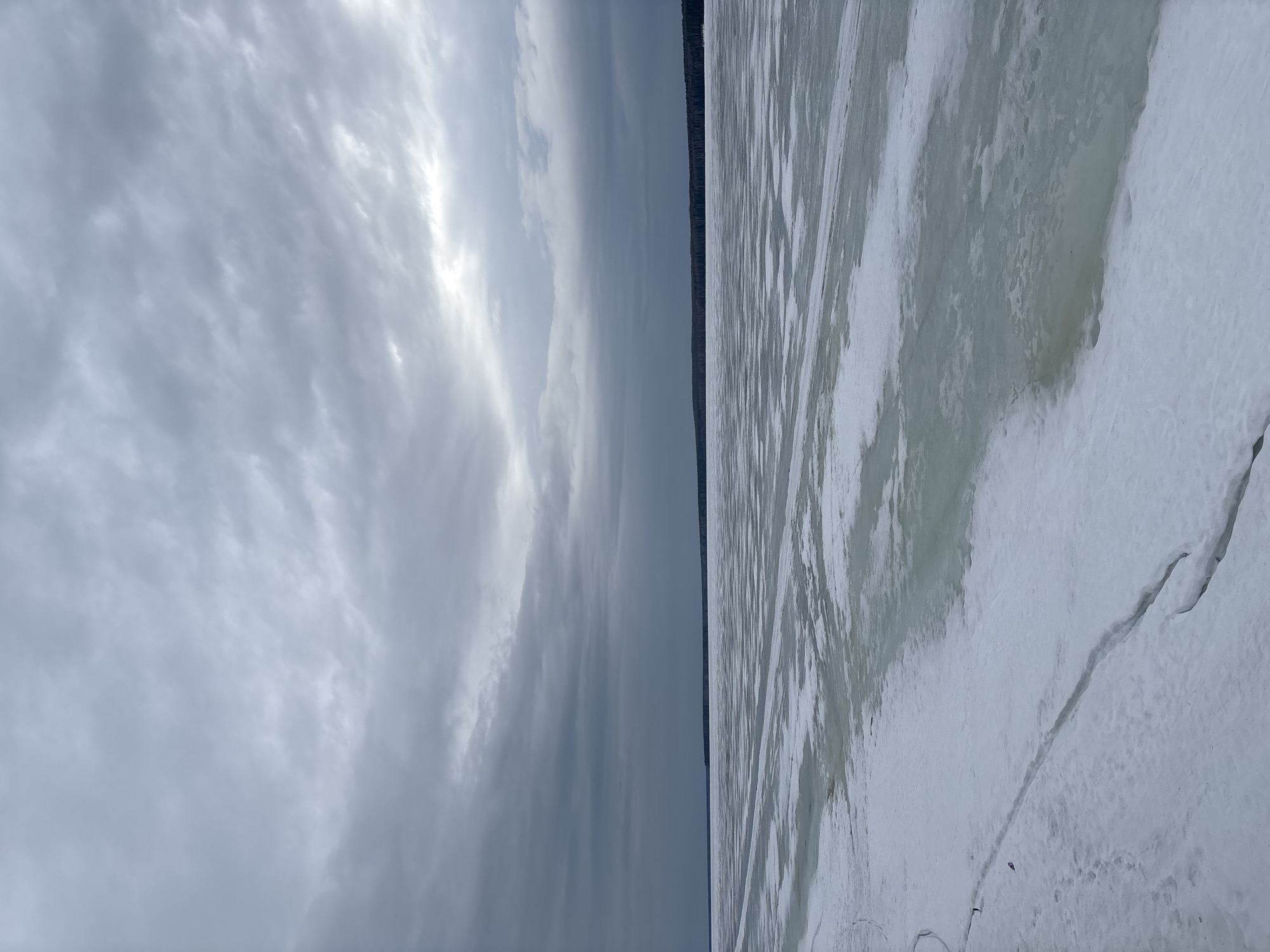 The image shows a landscape with a cloudy sky on the left and a partially frozen body of water (likely the Kama River) on the right. A thin line of trees is visible on the horizon separating the two. The ice is cracked and appears thin in some areas. The image was taken near Chaikovsky, Russia.