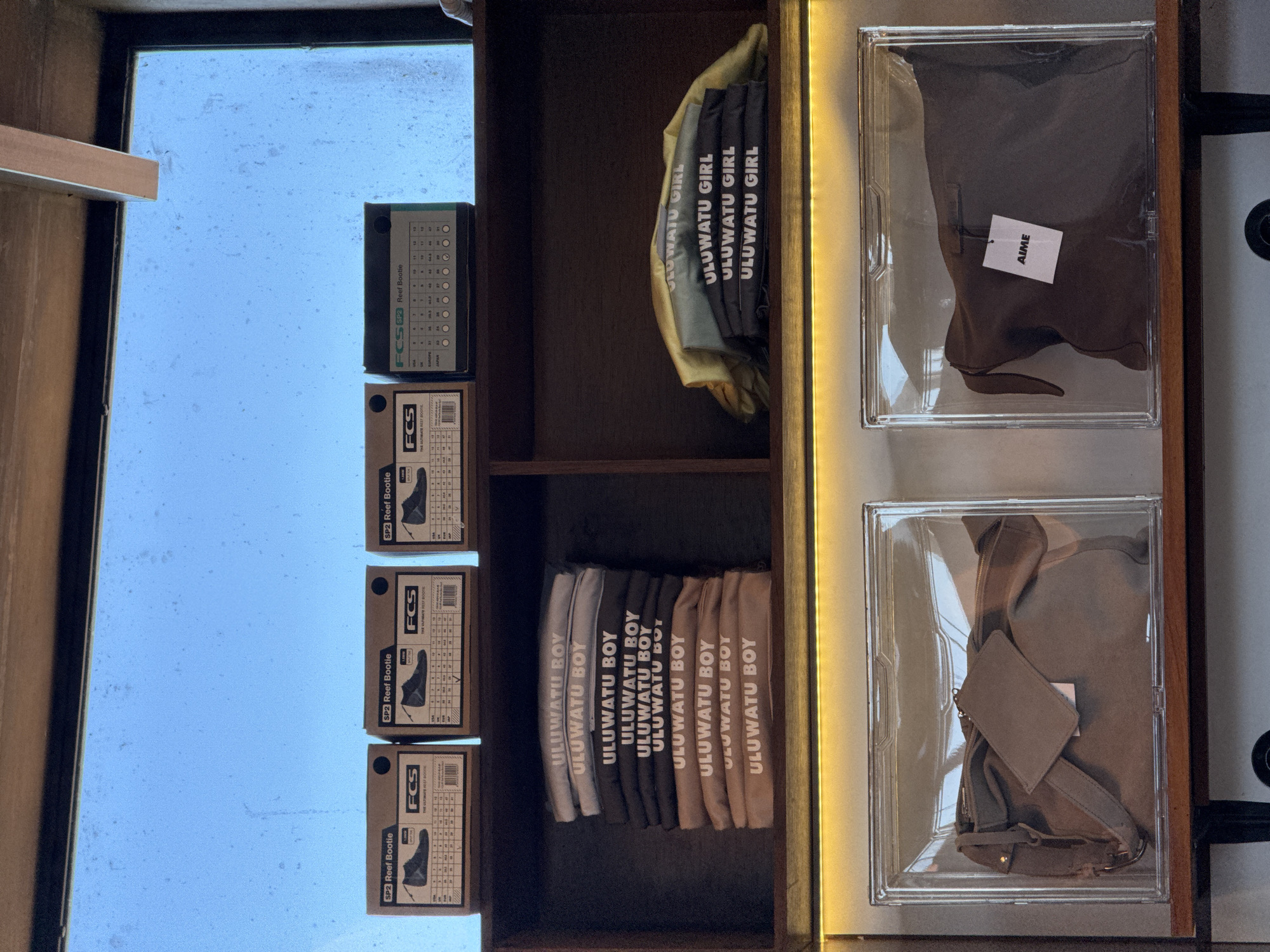 The image shows a display shelf with various items, likely in a retail setting. There are "FCS2 Reef Bootie" boxes stacked on the left, with sizing charts visible. In the middle section of the shelf, there are stacks of clothing items labeled "ULUWATU GIRL" and "ULUWATU BOY." On the right side of the shelf, there are two clear display cases containing folded clothing items, one labeled "AIME." The setting could be a surf shop or boutique, considering the items. The location, Kuta Selatan, Indonesia, can be inferred from the "ULUWATU" branding, as Uluwatu is a popular surfing destination in that region.
