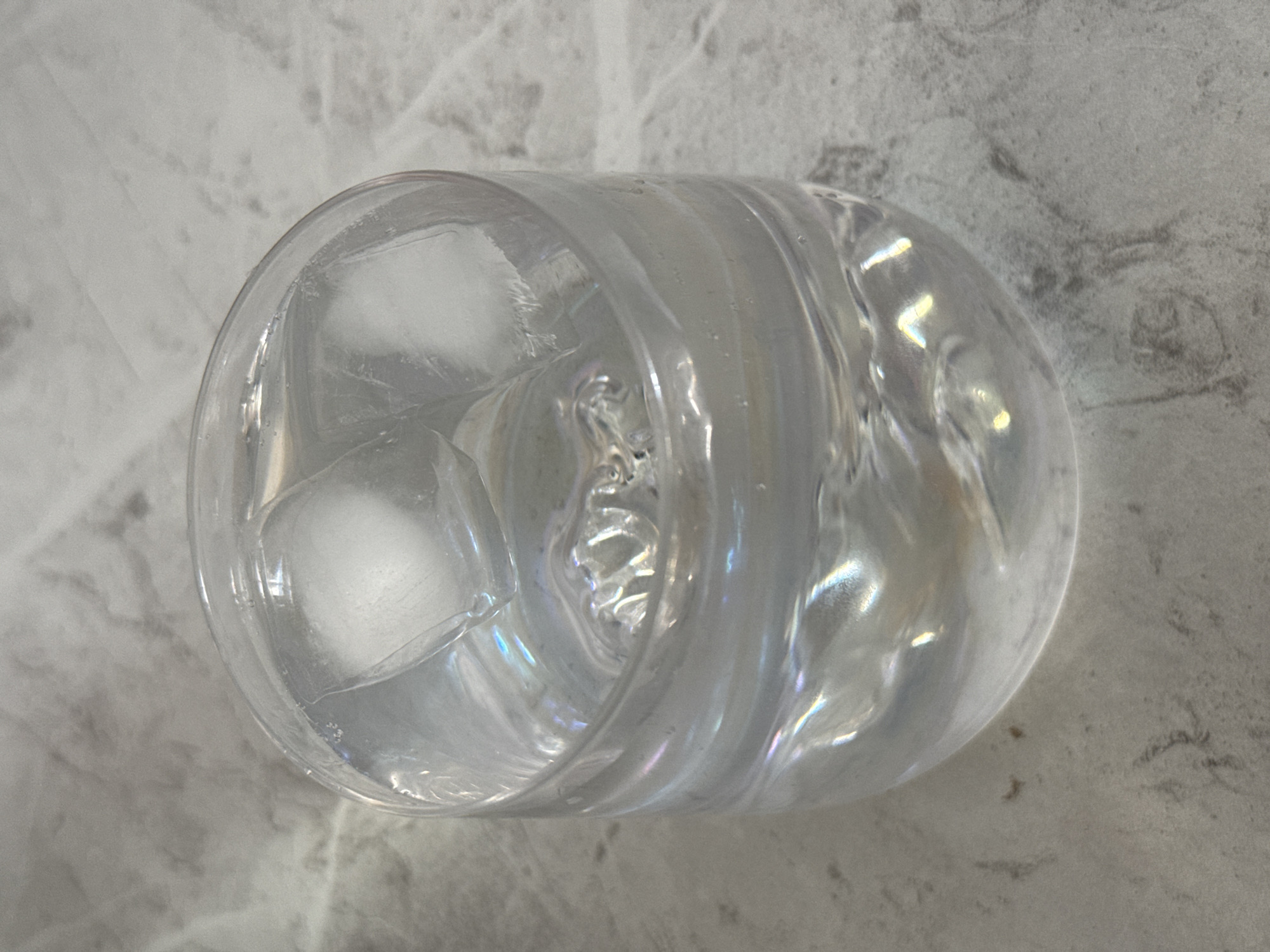 A close-up, high-angle shot shows a clear glass tumbler filled with water and ice cubes. The glass is placed on a light gray, textured surface that appears to be a marble countertop. The water is clear and reflects light, creating iridescent patterns on the surface of the glass and the water's surface. Three ice cubes are visible in the glass, with two partially submerged and one floating on top. The background is out of focus, but the texture of the countertop is visible. The scene is static and there are no people or animals present. The image does not contain any information about the location in Lyubertsy, Russia.
