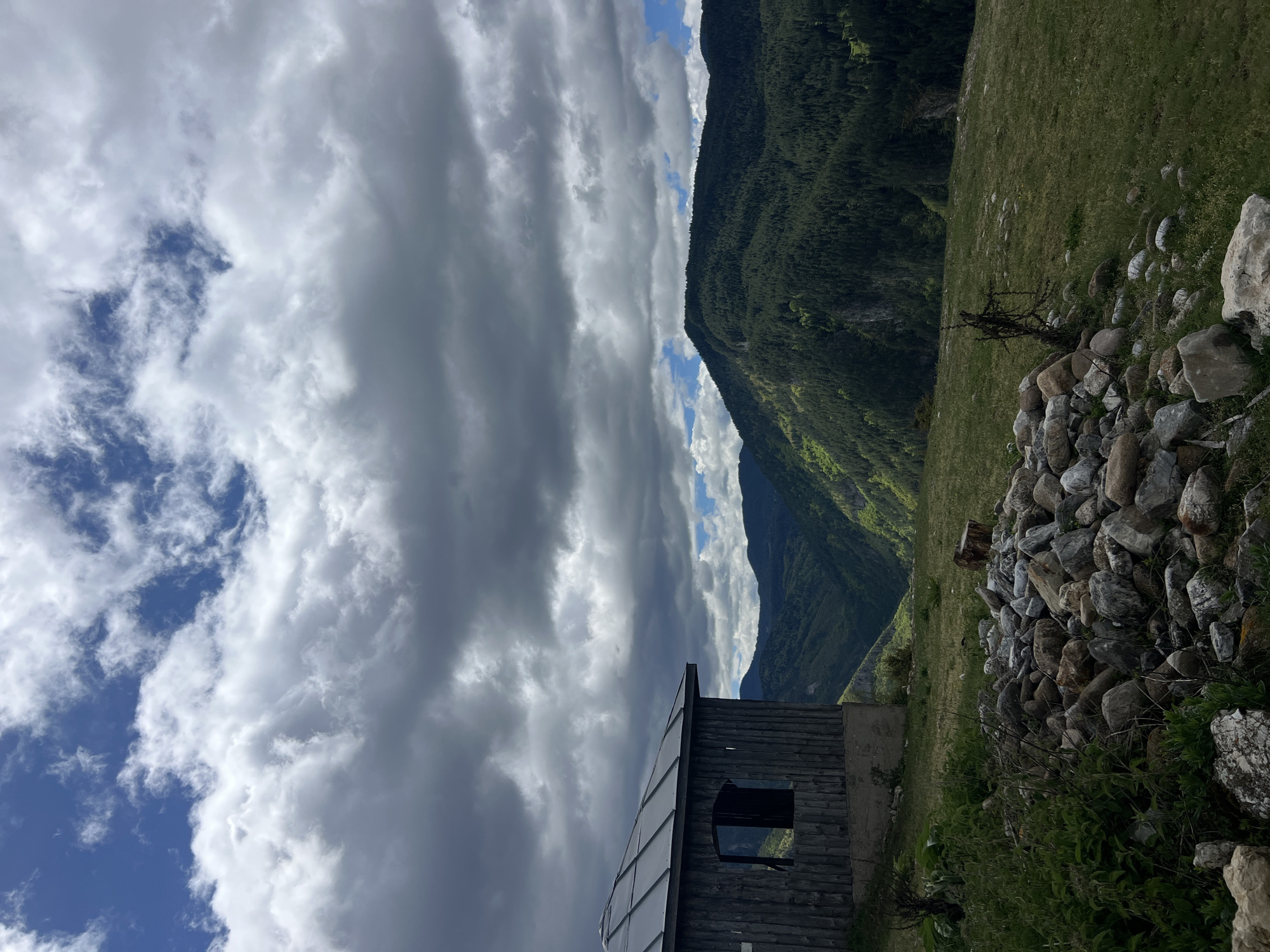 The image depicts a mountainous landscape in Giovren, Bulgaria. A small wooden structure stands in the foreground with a window facing the viewer. To the right of the structure, a pile of rocks is arranged on a grassy slope. In the background, a forested mountain rises, partially obscured by dramatic cloud formations. The sky features a mix of dark gray clouds and patches of blue.