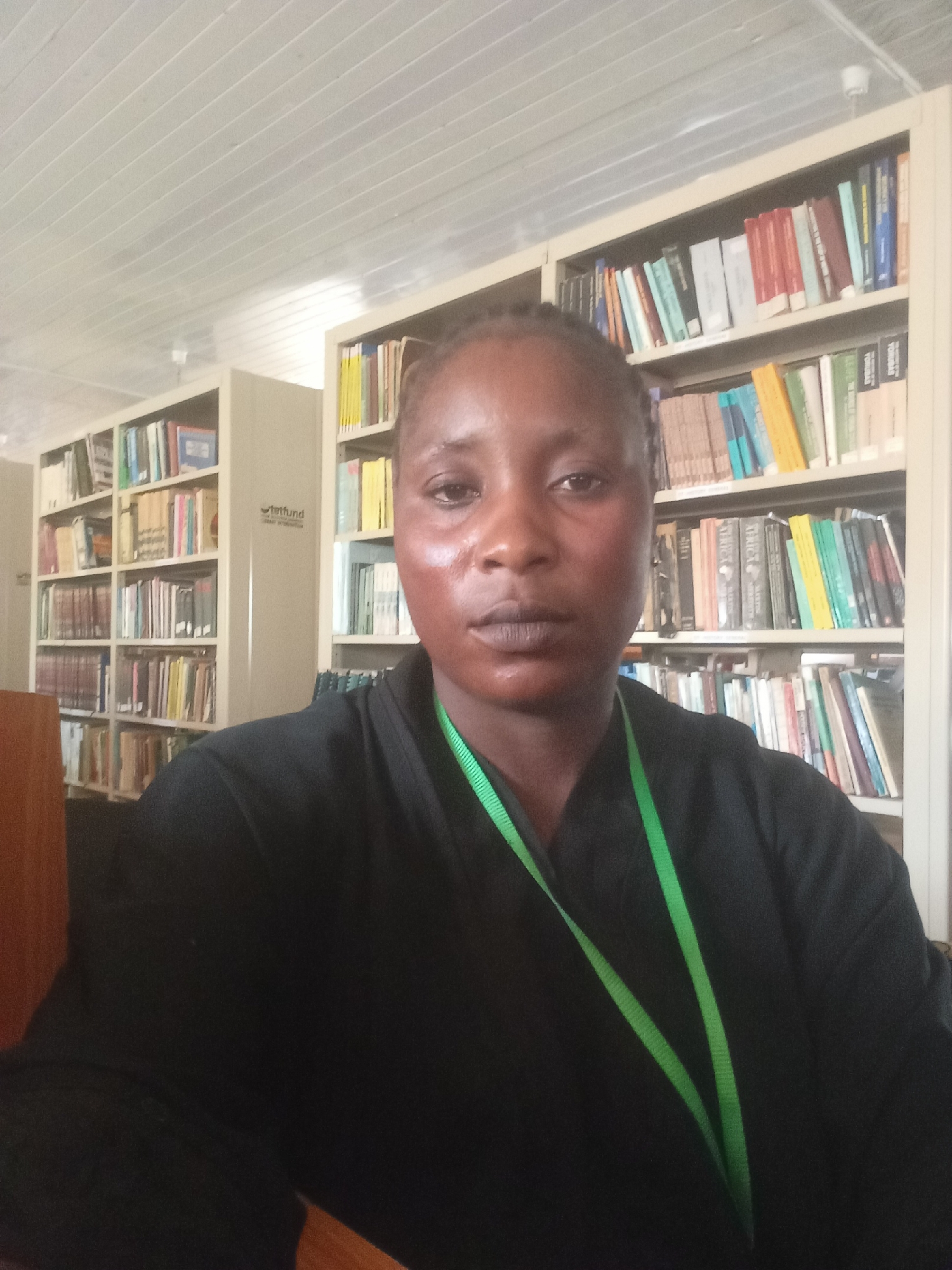 The image shows a person with brown skin, short, dark hair pulled back, wearing a black shirt and a green lanyard. The person is facing the camera with a neutral expression. Behind the person are bookshelves filled with books. The location appears to be a library. Akwanga, Nigeria is mentioned in the request.