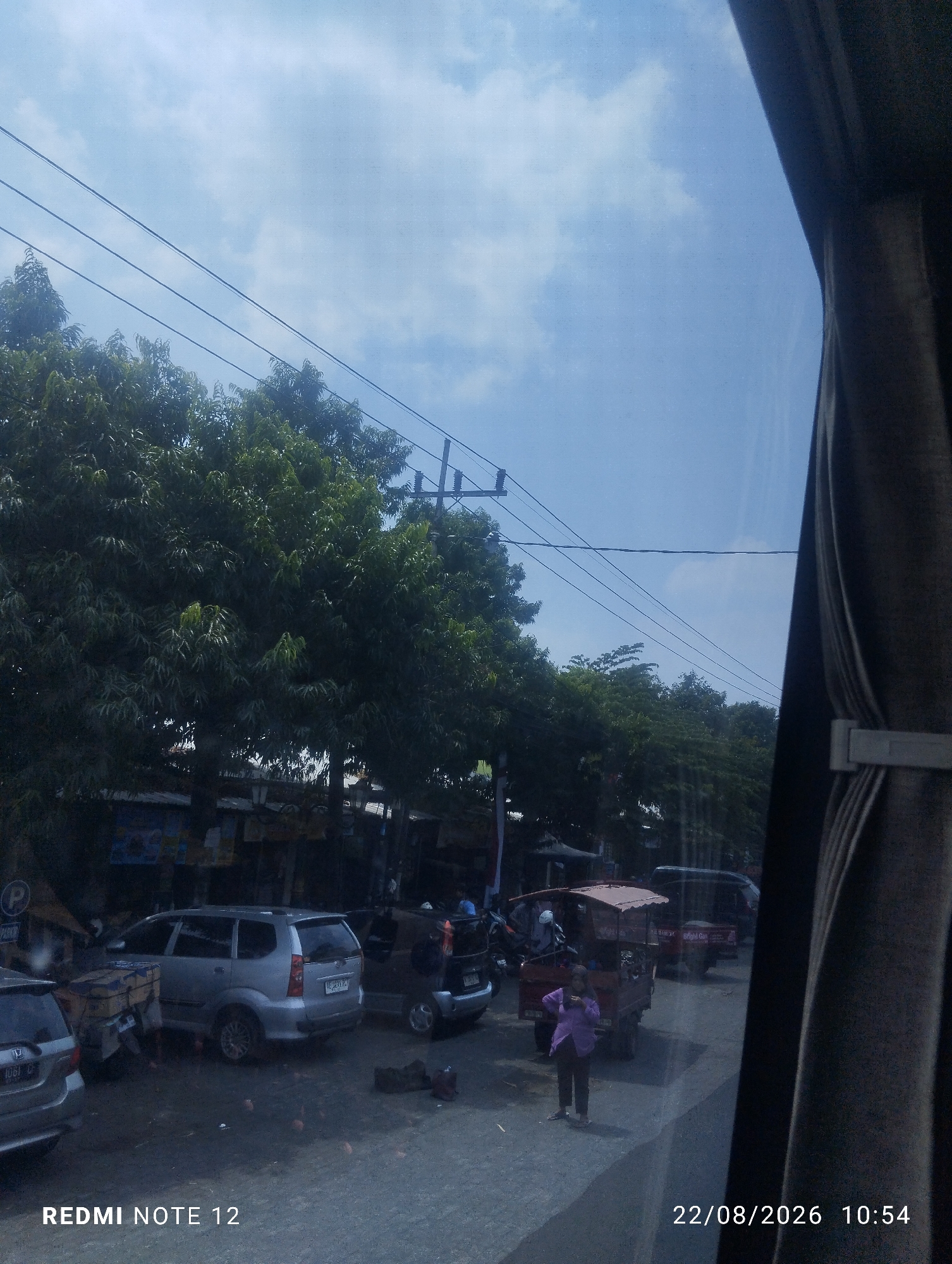 The image depicts a street scene in Maospati, Indonesia, on August 22, 2026, at 10:54 AM. The perspective is from inside a vehicle, looking out through a window partially obscured by a dark curtain on the right side. The sky is bright blue with scattered clouds. Several power lines stretch across the upper portion of the frame.
In the mid-ground, a line of trees with dense green foliage runs along the side of the road. Below the trees, several vehicles are parked or in motion. A silver minivan is prominently visible, facing right. To its left, a parked silver car is partially visible. Further down the road, a red cart with a canopy is positioned, and a person in a purple top and dark pants stands near it. Other vehicles, including motorcycles and potentially another van, are also present in the background.
The ground appears to be asphalt, with some debris and scattered items visible. The overall impression is that of a typical street scene in a developed area, possibly a market or commercial zone, given the presence of parked vehicles and the type of stalls visible beneath the trees. The timestamp and "REDMI NOTE 12" are overlaid on the bottom of the image.
