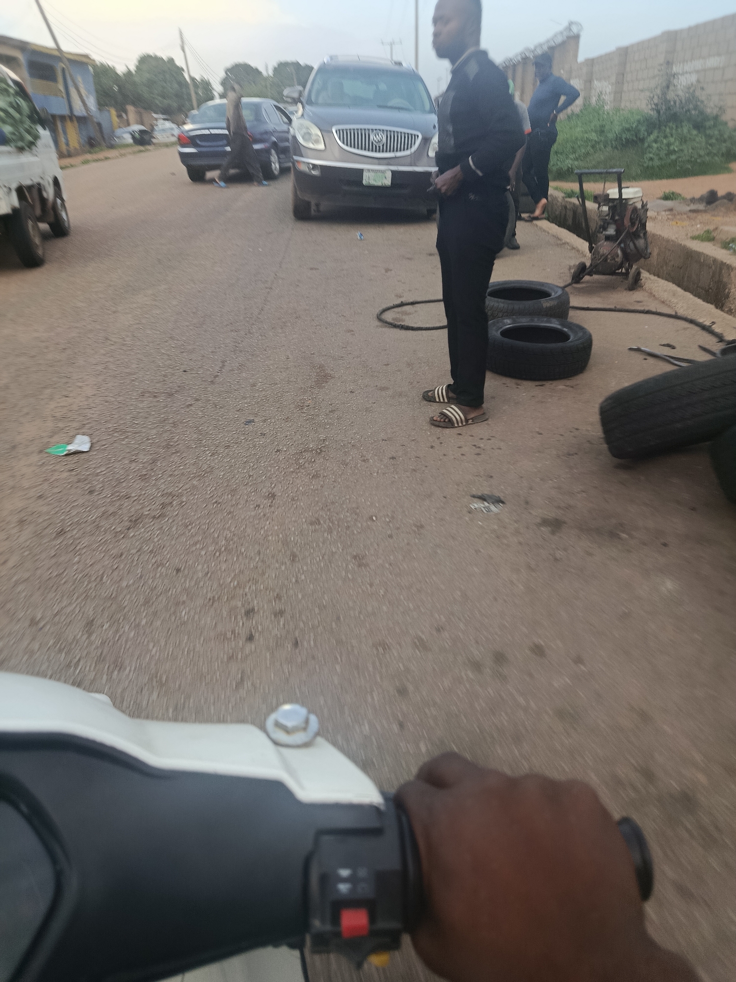 A man in a black suit and sandals stands on a paved road near several tires. Behind him, a car, likely a Buick, is parked. Another man in dark clothing walks away in the background, and a small, wheeled machine, possibly a generator or compressor, sits on the side of the road. The scene appears to be in a semi-urban or rural area, with buildings and vegetation visible in the distance. The image quality is somewhat blurry, suggesting it was taken while in motion, possibly from a motorcycle. This is likely Rafin Par, Nigeria.