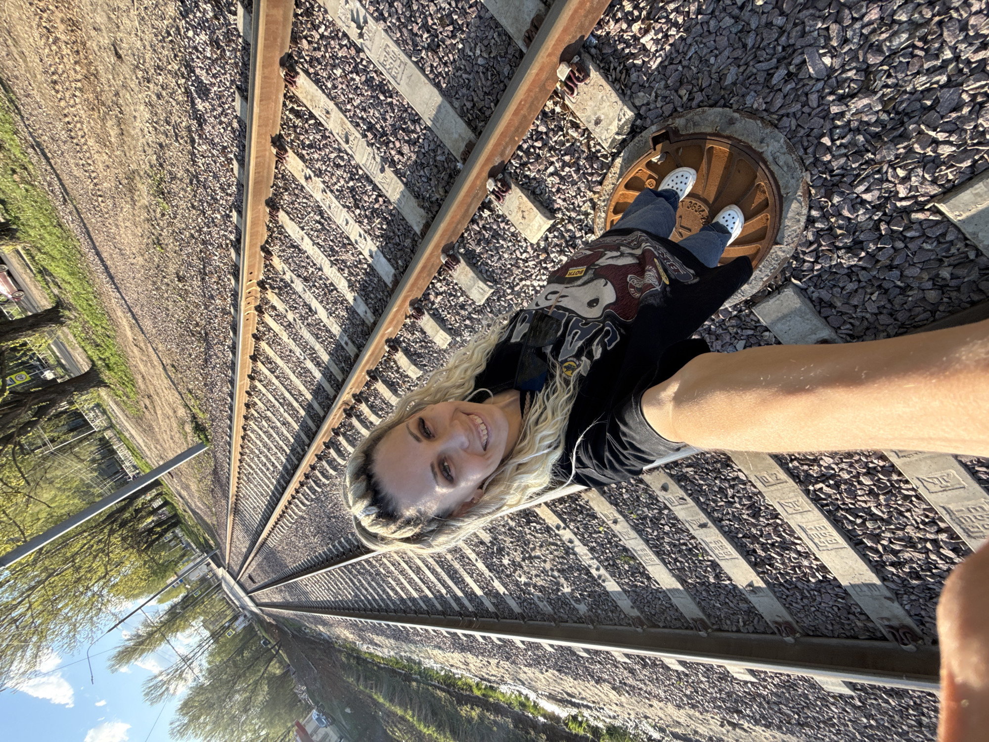 A woman is positioned on a set of railroad tracks. She is standing on a manhole cover between the rails. The location appears to be in Rusanovo, Russia. She is wearing white shoes, blue jeans, and a black t-shirt. The railroad tracks are surrounded by gravel. Trees are visible on the left side of the image. The sky is partially visible in the top left corner.